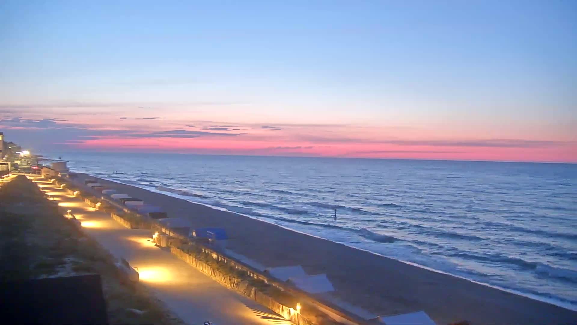 A pink and blue sunrise illuminates a beach with a lighted walkway lined with structures, and gentle waves roll onto the shore.