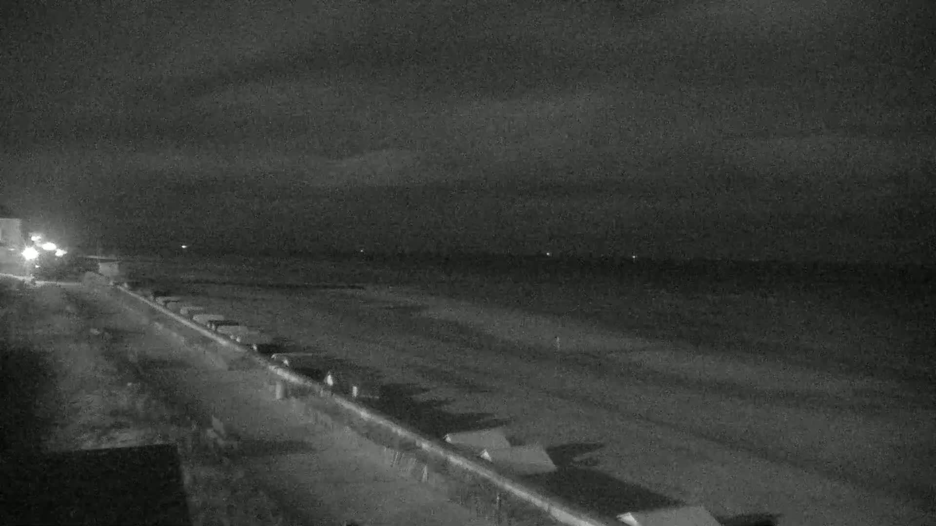 A dark, nighttime view of a beach with a roadway and small structures along the shoreline under a cloudy sky.