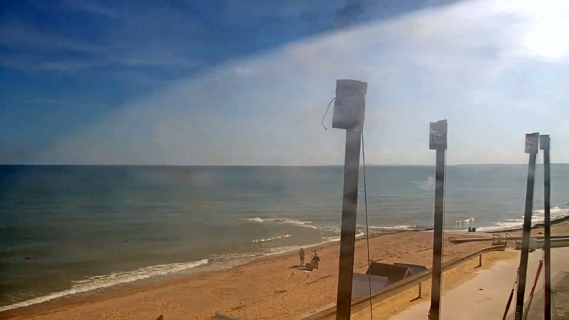 A sandy beach with a few people on it meets a calm, light-blue ocean under a mostly sunny sky with some hazy clouds.