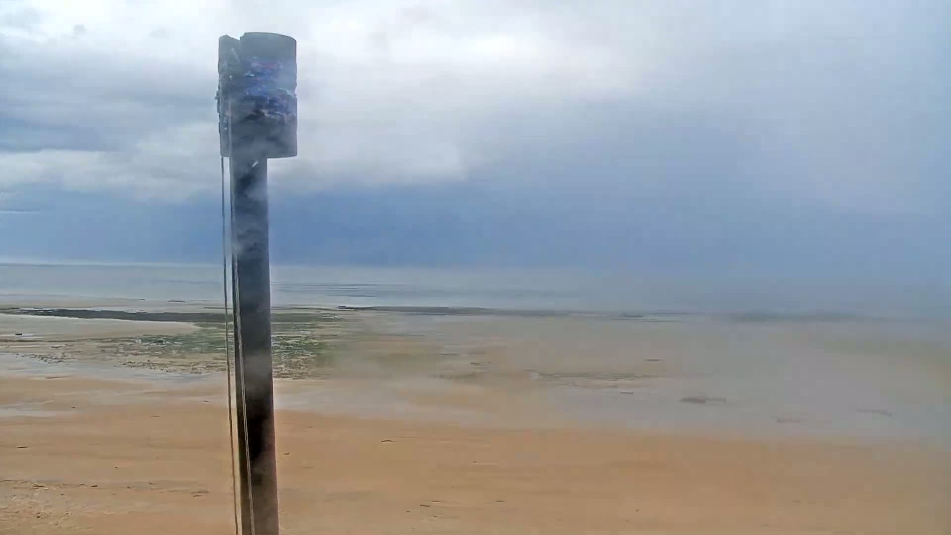 A tall, thin, dark pole stands on a sandy beach overlooking a hazy, overcast ocean.