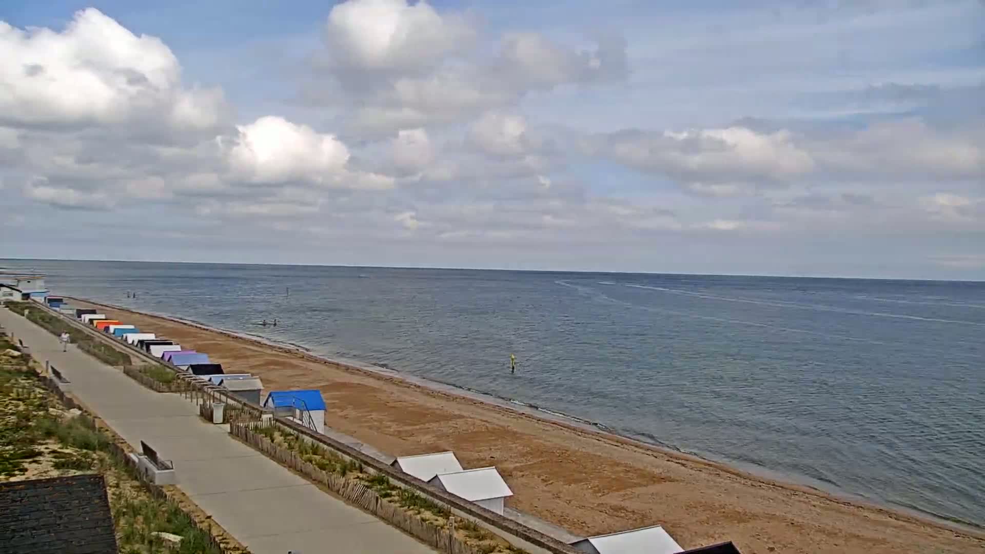 A sandy beach with colorful beach huts lines a paved walkway next to a calm sea under a partly cloudy sky.