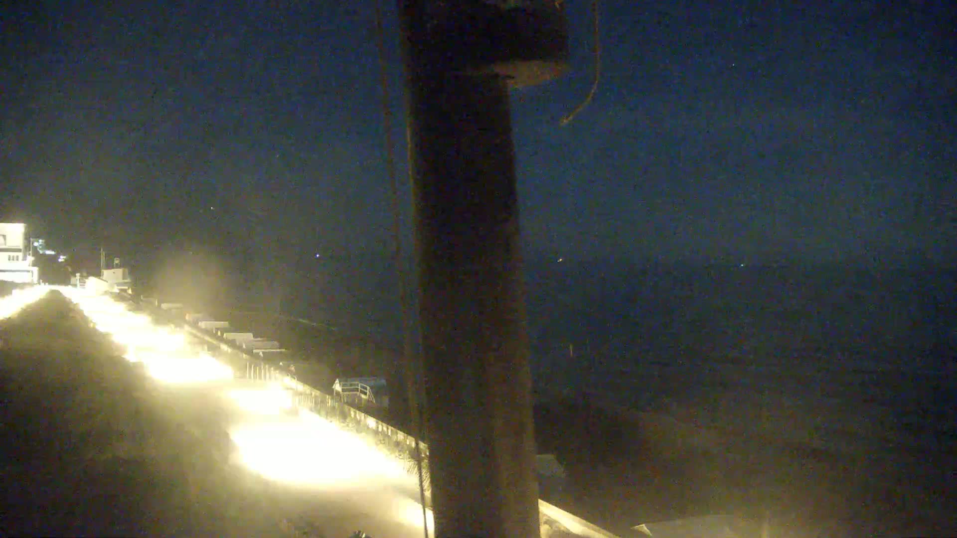 At night, a line of headlights from vehicles travels along a roadway, with a dark body of water visible in the background.