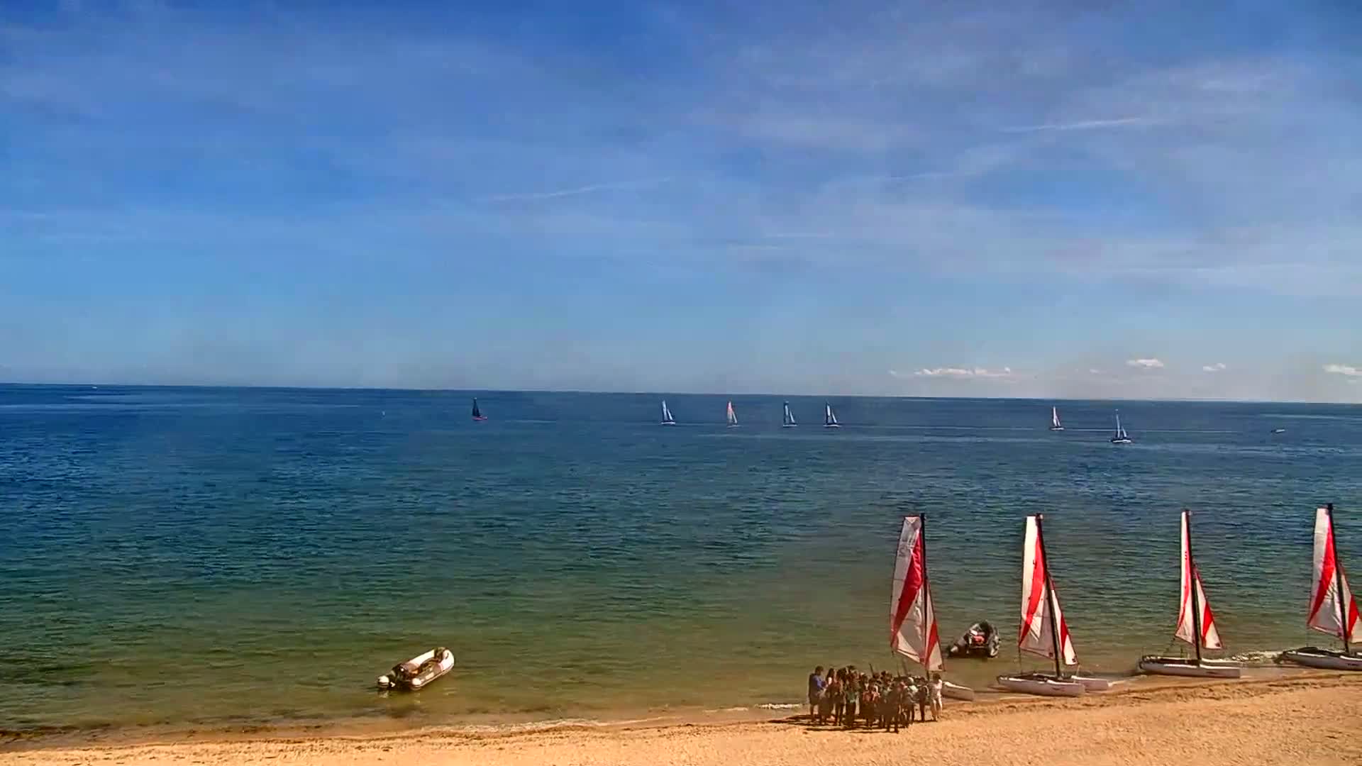 A sandy beach with several small sailboats on the shore and a few more sailing further out in a calm, sunny ocean.