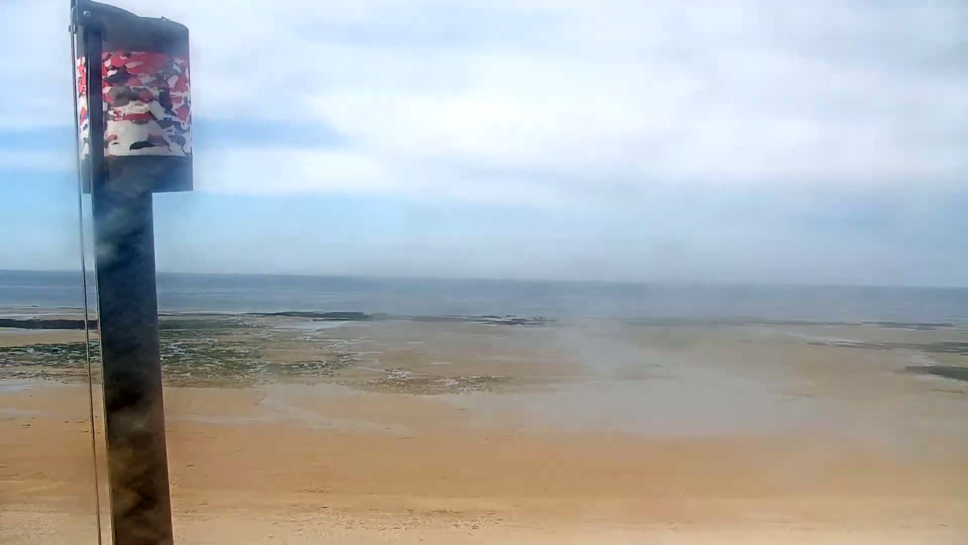 A sandy beach stretches to a calm ocean under a hazy sky, with a tall pole bearing a patterned cylindrical object in the foreground.