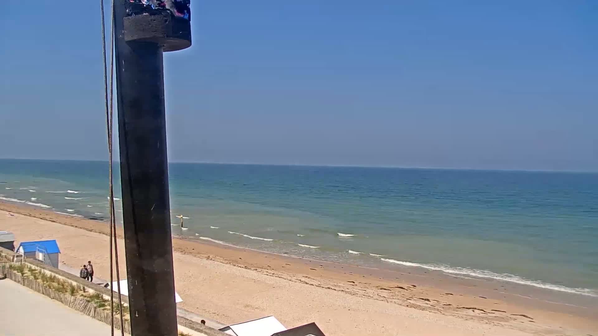 A sandy beach with gentle waves under a clear blue sky is viewed from behind a tall dark post.