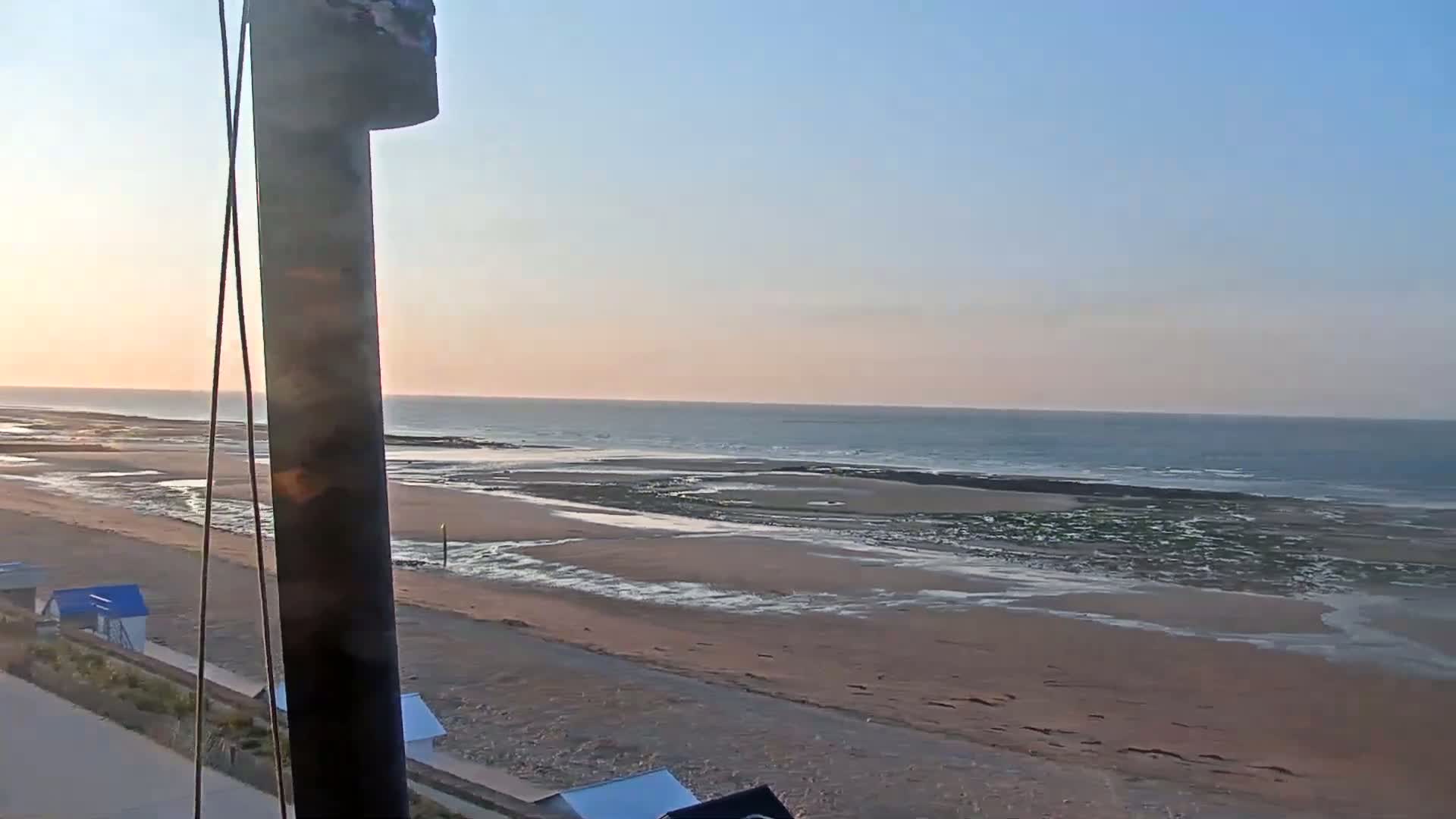 A sandy beach stretches towards a calm ocean under a clear, pale sunset sky.