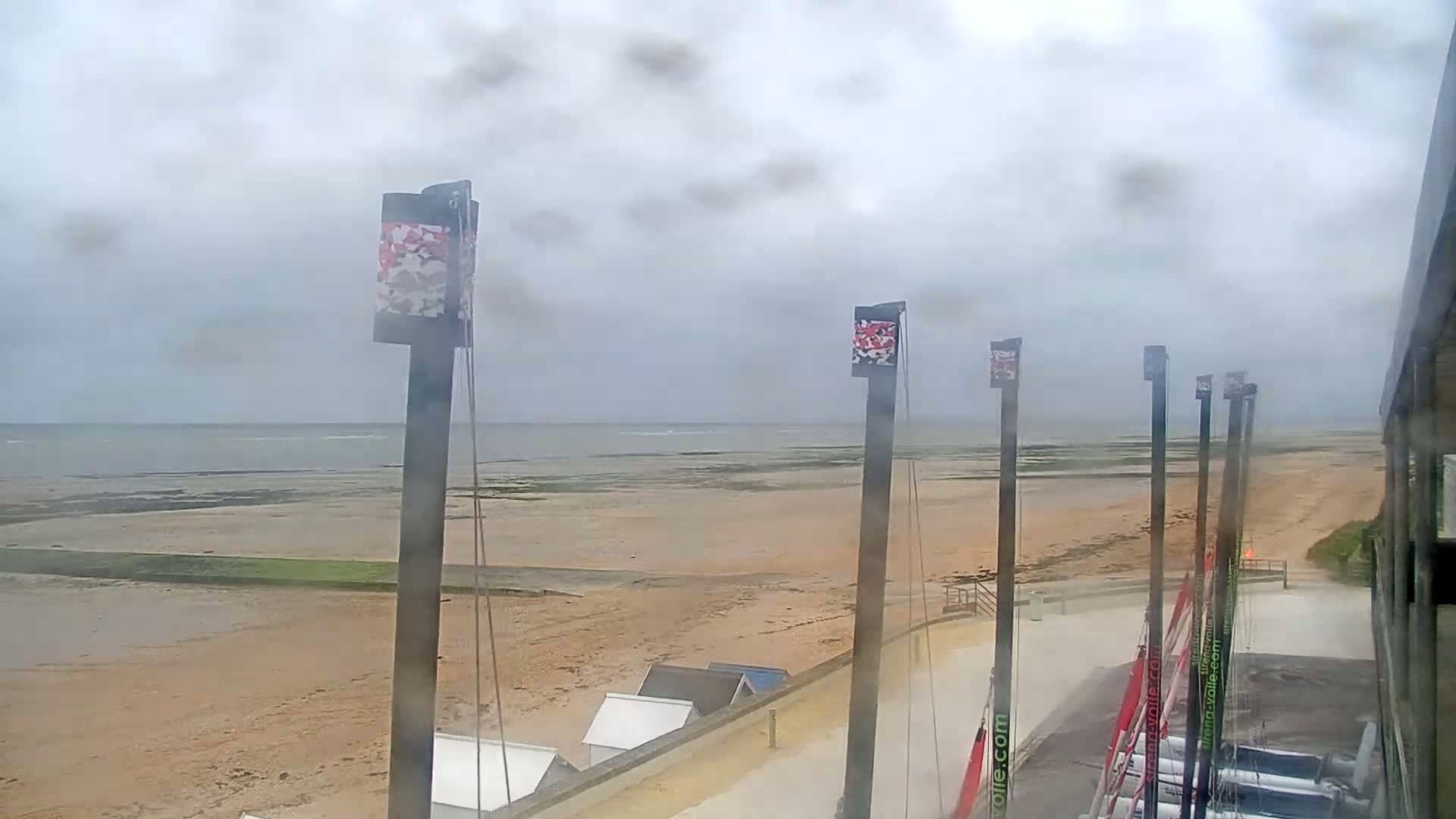 A sandy beach stretches to a calm sea under an overcast sky, with several tall posts bearing flags visible in the foreground.