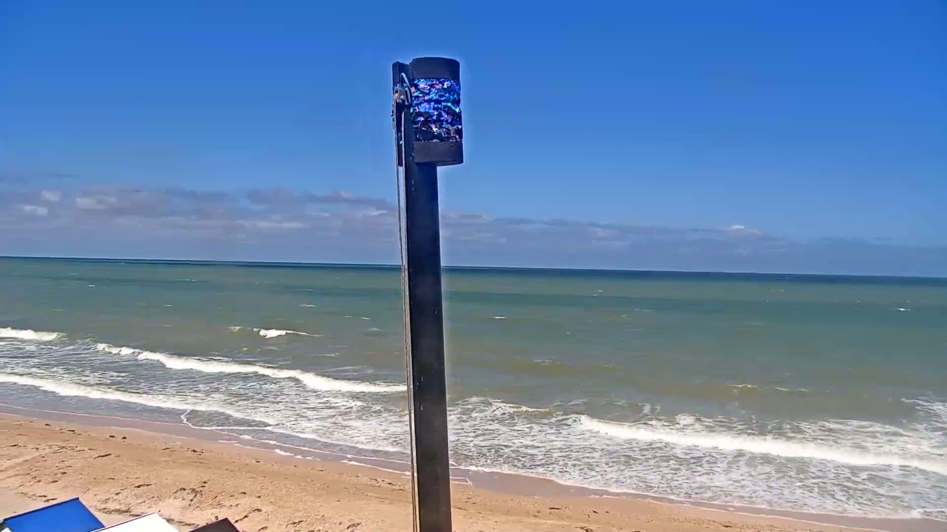 A tall, thin, dark pole with a decorative top stands on a sandy beach overlooking a wavy ocean under a mostly clear blue sky.