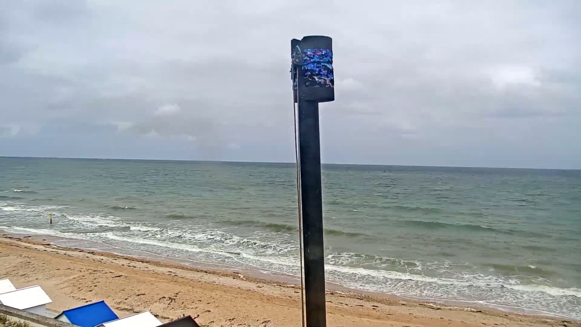 A tall, dark post stands in front of a sandy beach with gentle waves rolling onto the shore under a cloudy sky.