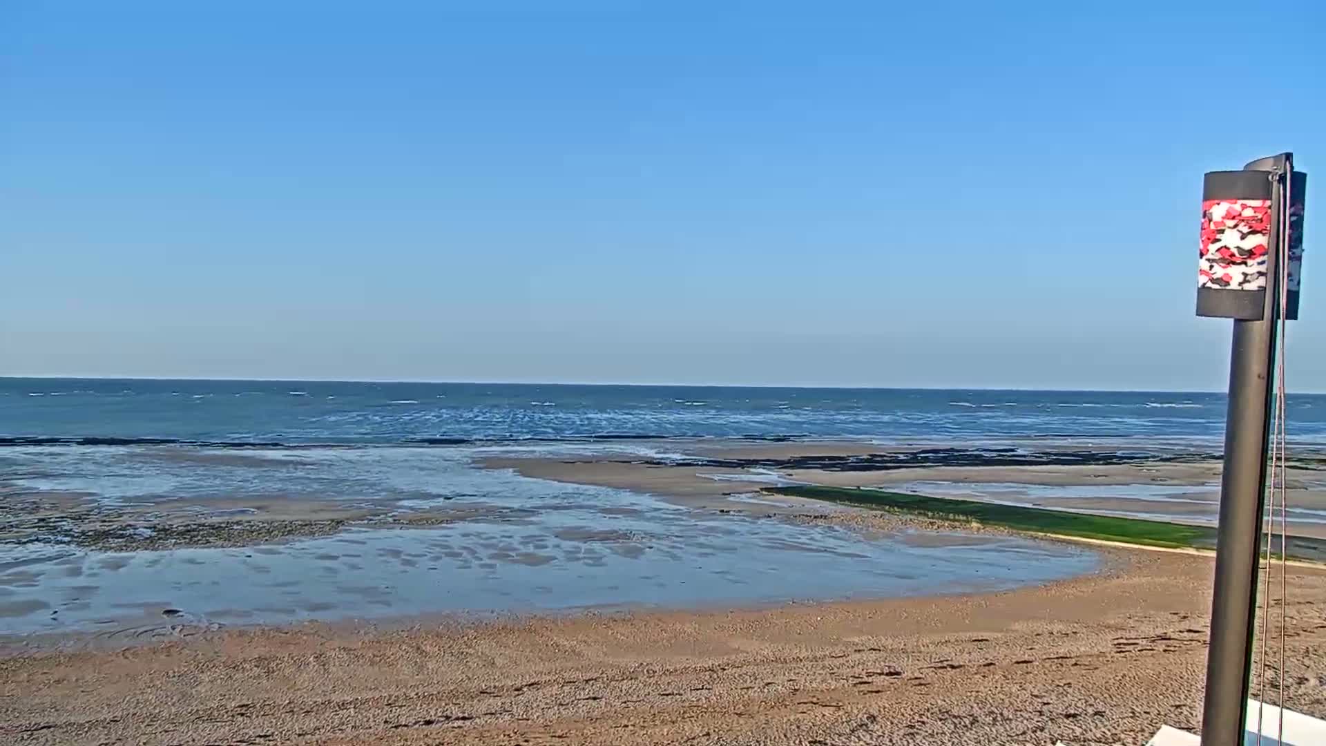 A sandy beach with shallow, receding water stretches toward a calm, blue ocean under a clear sky.