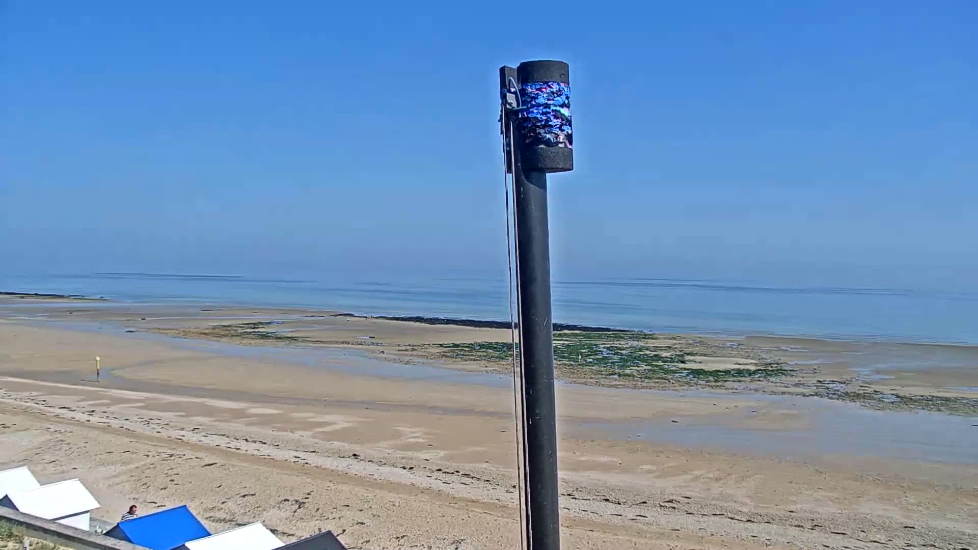 A tall pole with a cylindrical object on top stands in the foreground, overlooking a wide sandy beach and calm ocean under a clear blue sky.