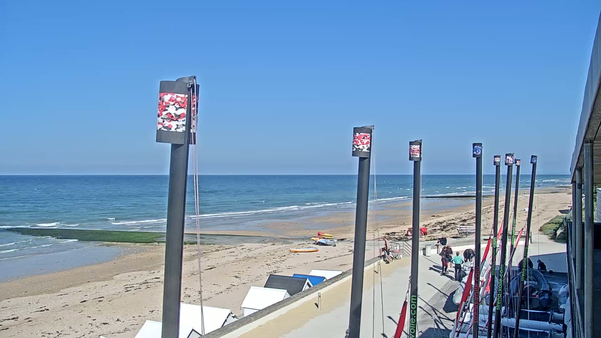 A sandy beach with calm ocean waves under a clear blue sky features several sailboats and beach equipment near a building.