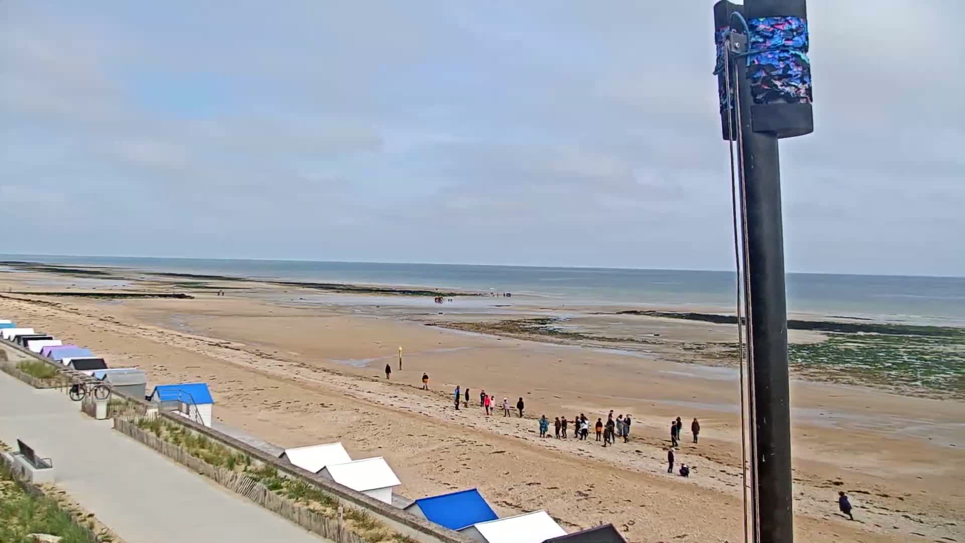 A sandy beach with small beach huts and a group of people stretches toward a calm, light-blue ocean under a mostly cloudy sky.