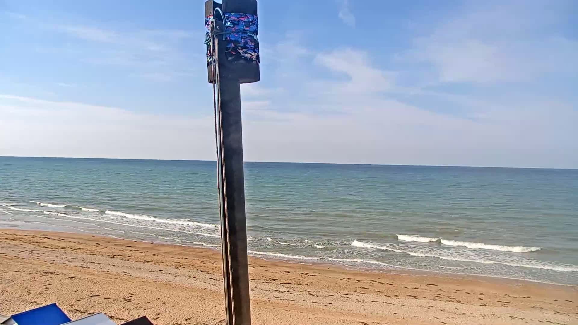 A sandy beach with gentle waves on a calm, sunny day is viewed from behind a dark pole with colorful cloth wrapped around its top.