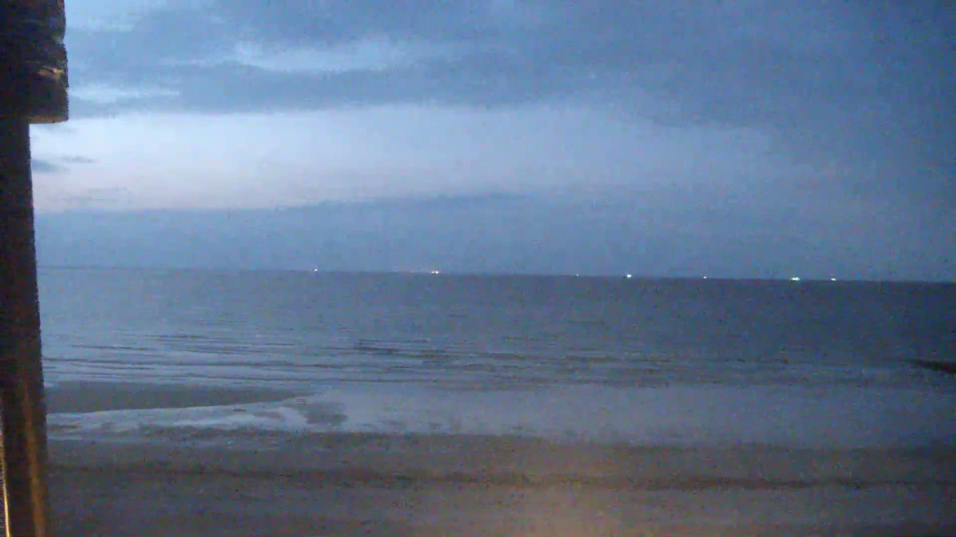A calm, dark-blue ocean meets a light-brown sandy beach under a cloudy, dusky sky.