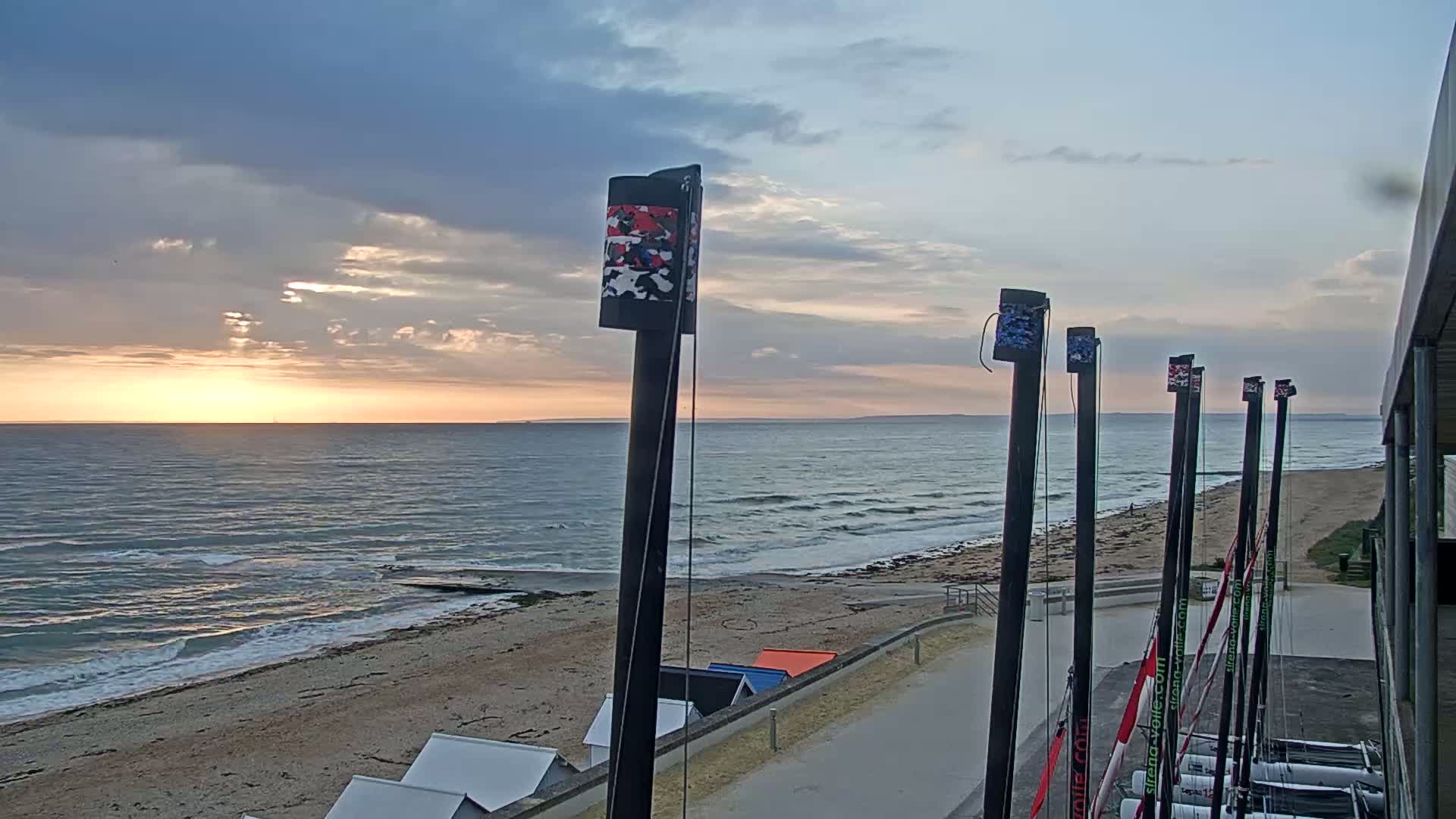 A beach with calm waves at sunset, several tall masts are in the foreground.