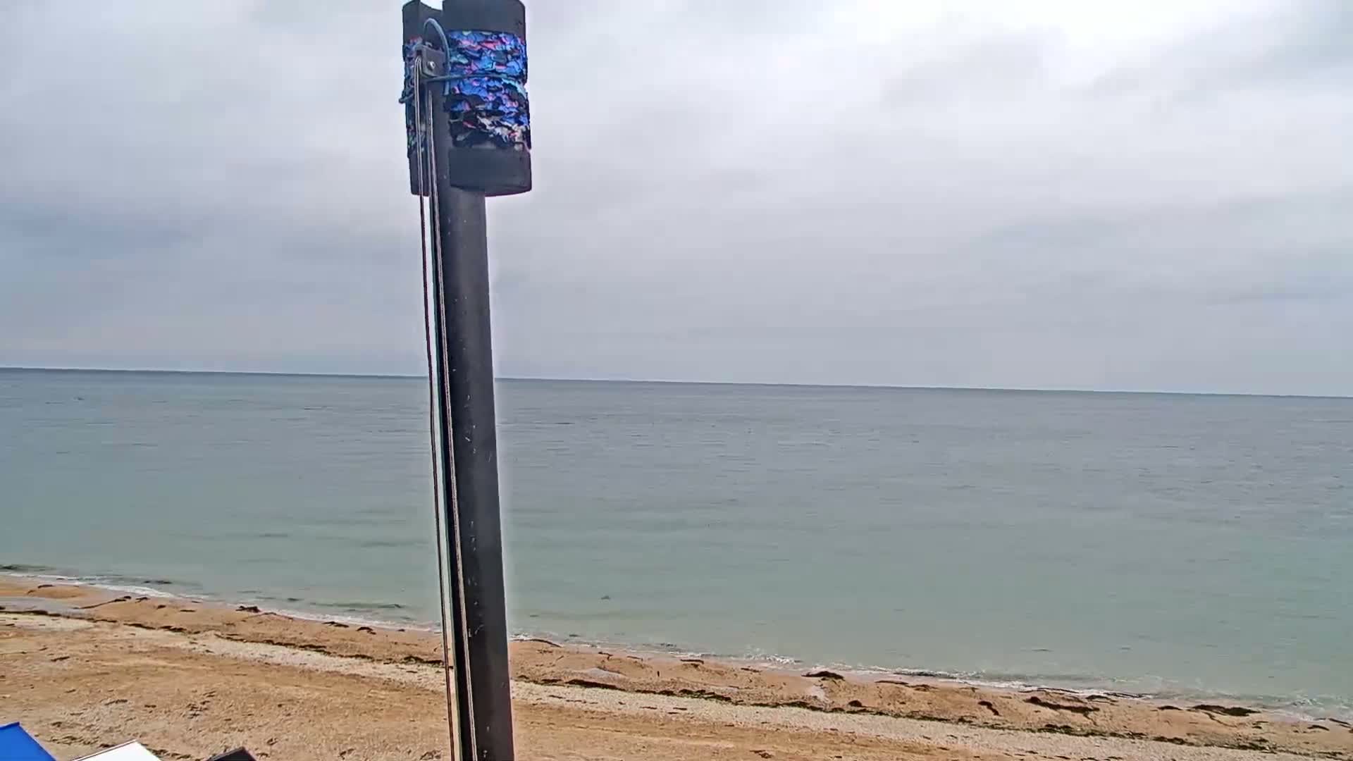 A tall, dark pole with a patterned cover stands in the foreground, overlooking a calm, gray-blue sea and a sandy beach under a cloudy sky.
