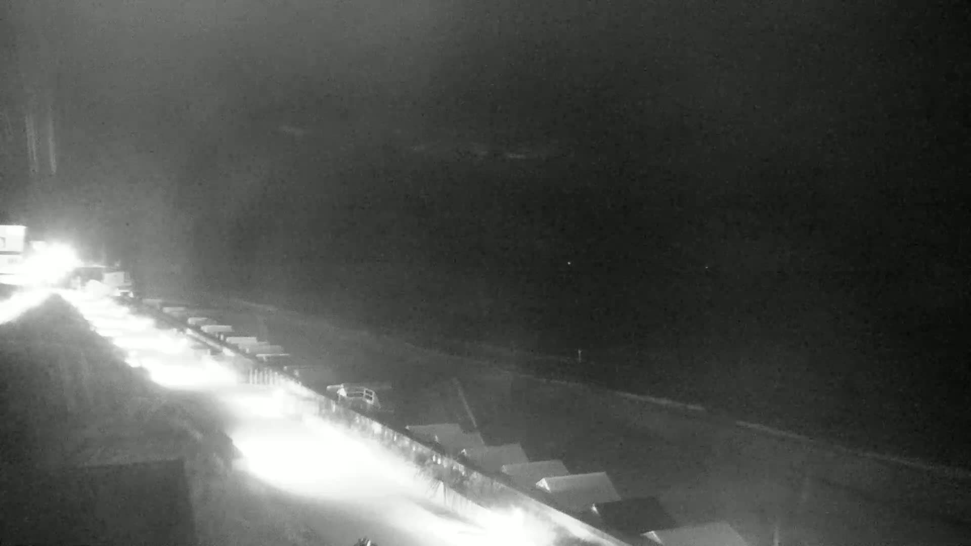 A nighttime, snowy scene shows a road with vehicles driving past a snow-covered area with buildings.