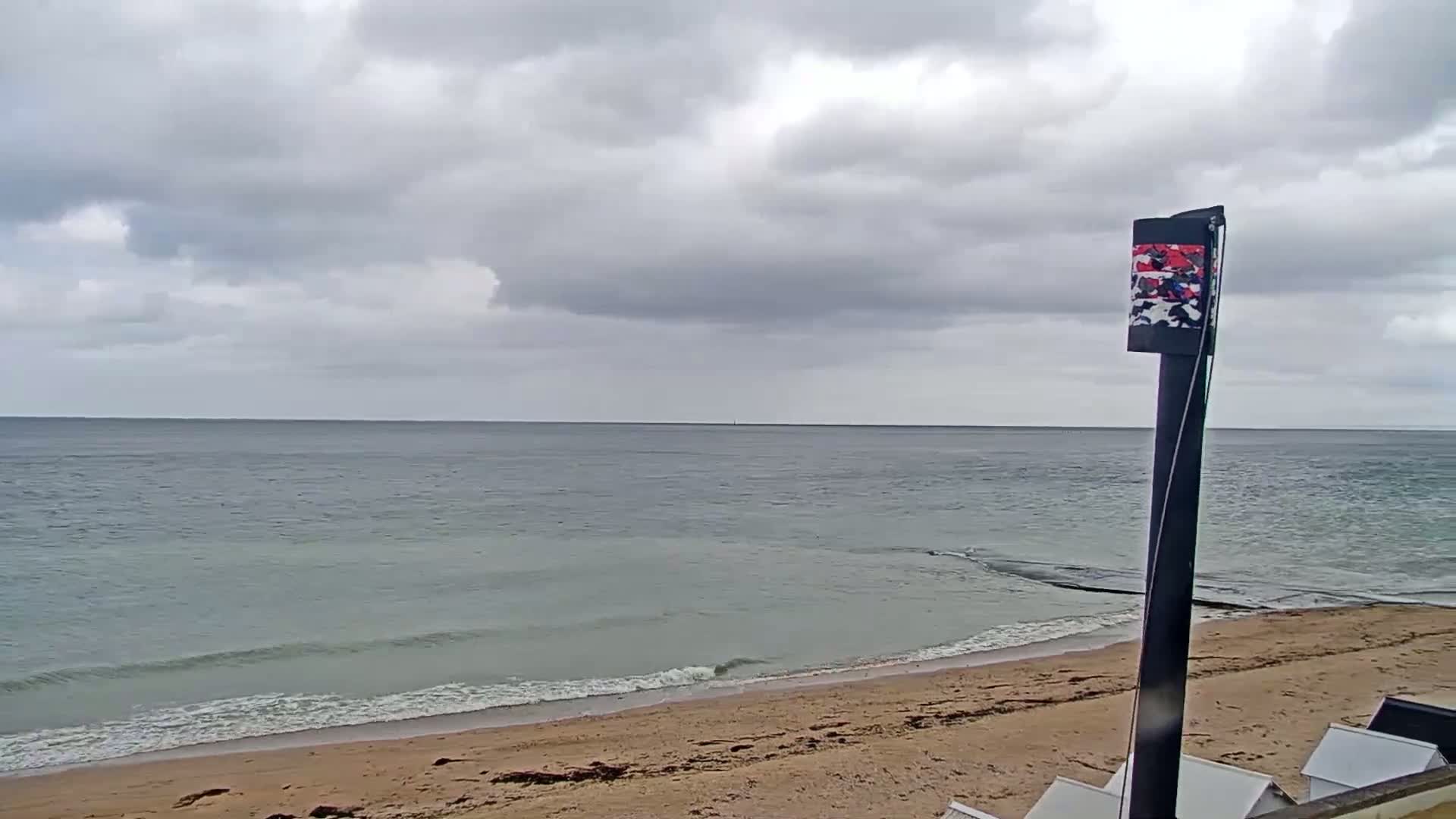 A calm, grey-sky day shows a sandy beach with gentle waves lapping the shore, a tall black post stands near the beach.