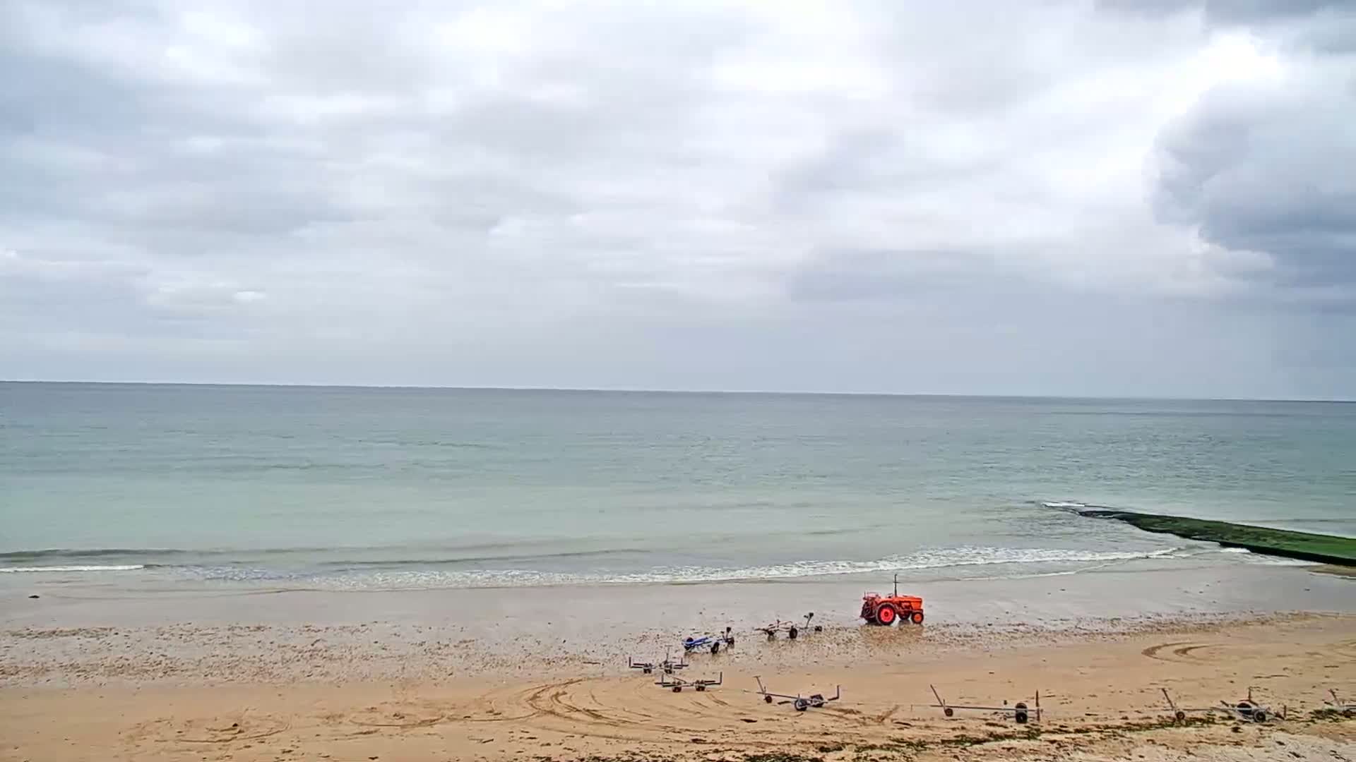 A tractor and several small trailers sit on a sandy beach under an overcast sky, next to a calm, light-green ocean.