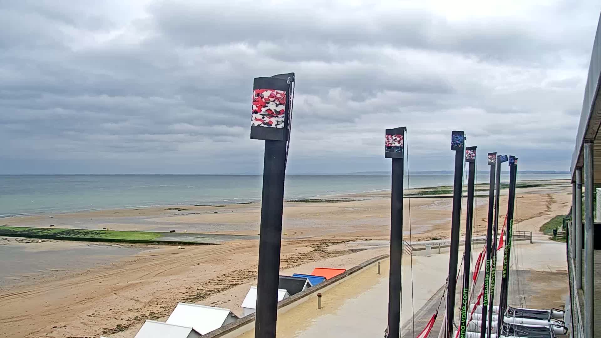 A sandy beach stretches to a calm, light-blue sea under a cloudy sky, with several tall masts bearing colorful flags standing near a row of beach huts.