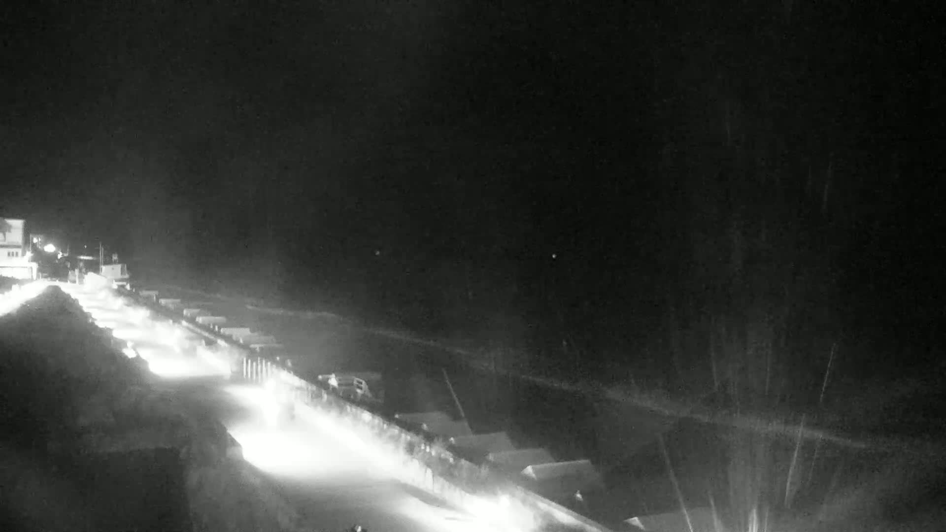 A snow-covered road at night is illuminated by headlights of vehicles traveling along it, with buildings visible in the distance under a dark, snowy sky.