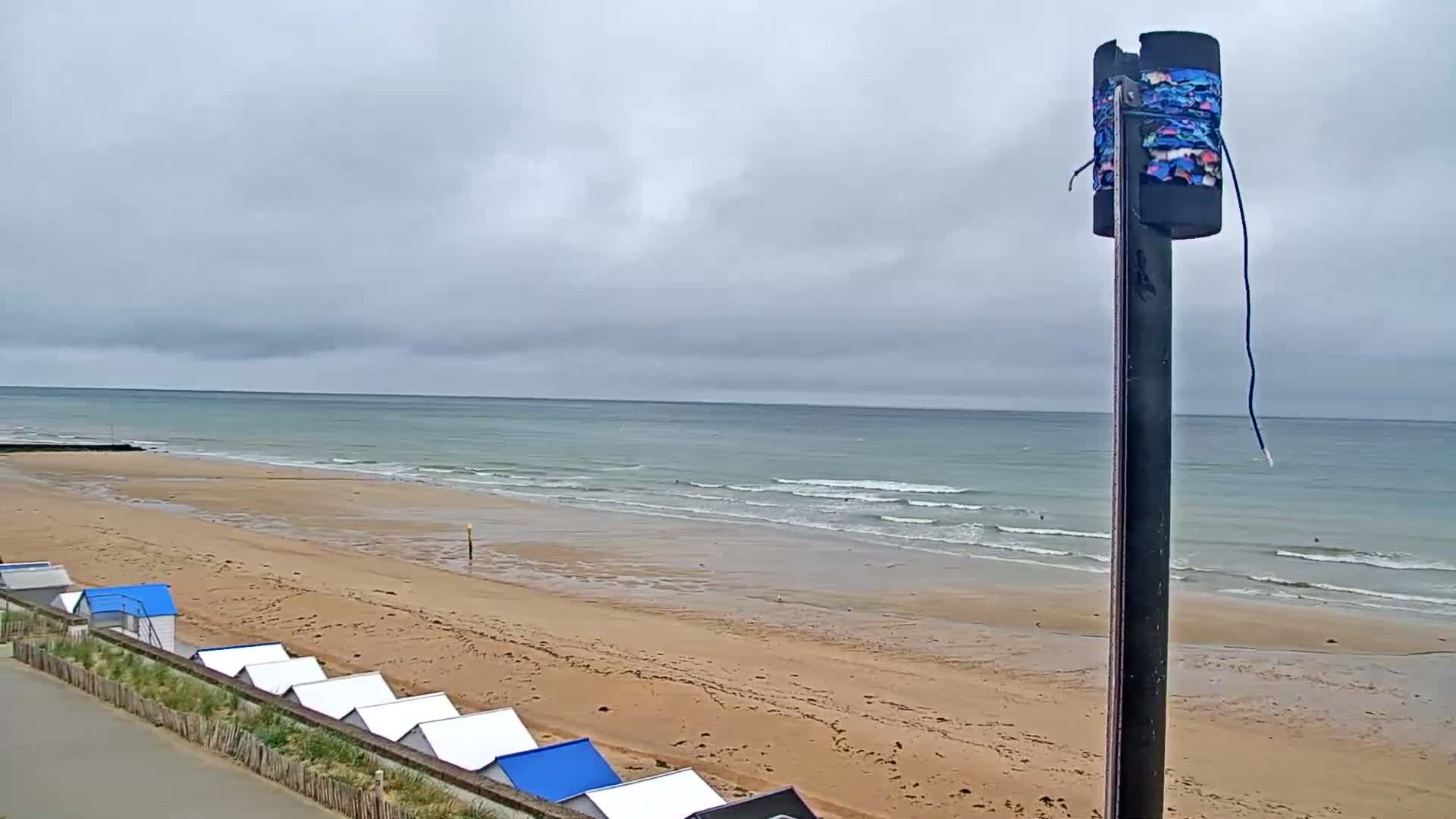 A sandy beach with small, blue-roofed beach huts stretches along a calm, ocean under a cloudy sky.