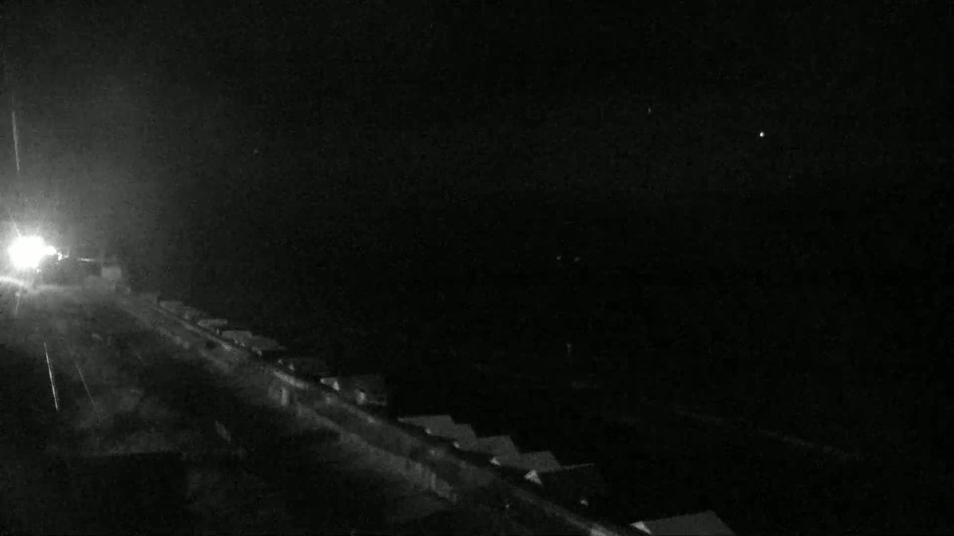 A nighttime, low-light view shows a road with buildings to the right and a bright light source in the distance on the left.