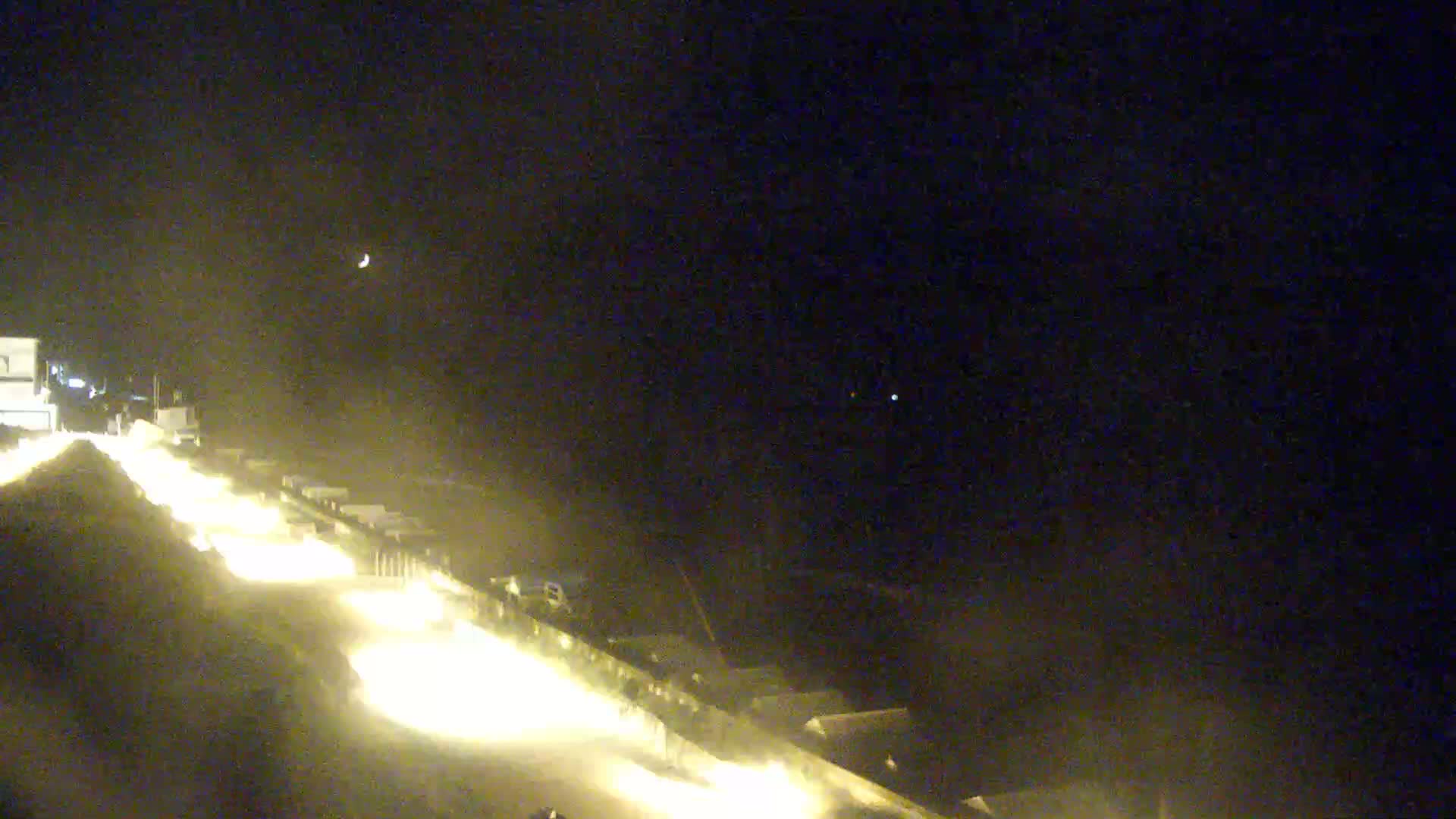 At night, under a crescent moon, vehicle headlights illuminate a roadway and surrounding structures.