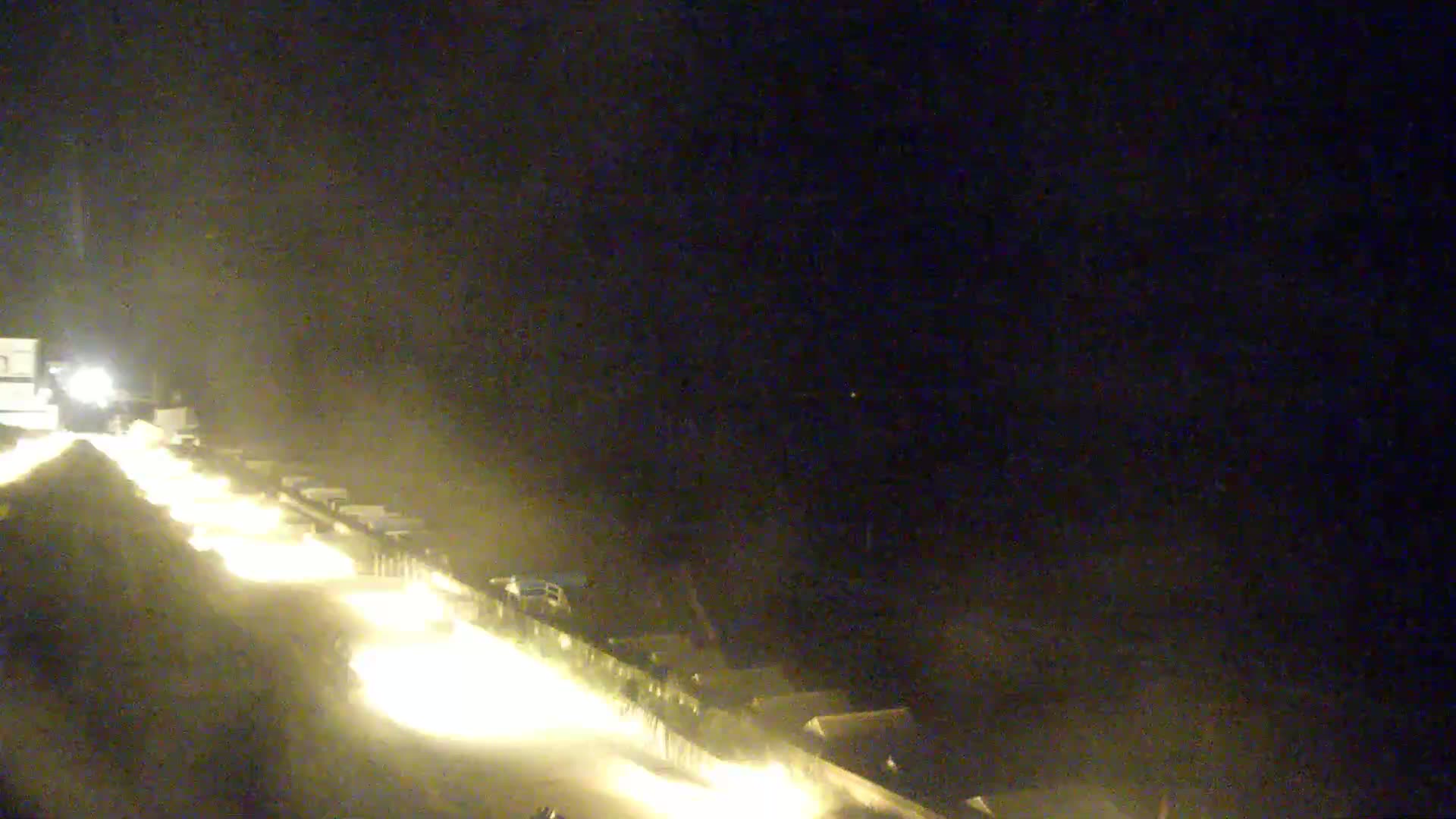 A nighttime, dimly lit scene shows a line of vehicles traveling along a roadway, their headlights illuminating the road ahead.