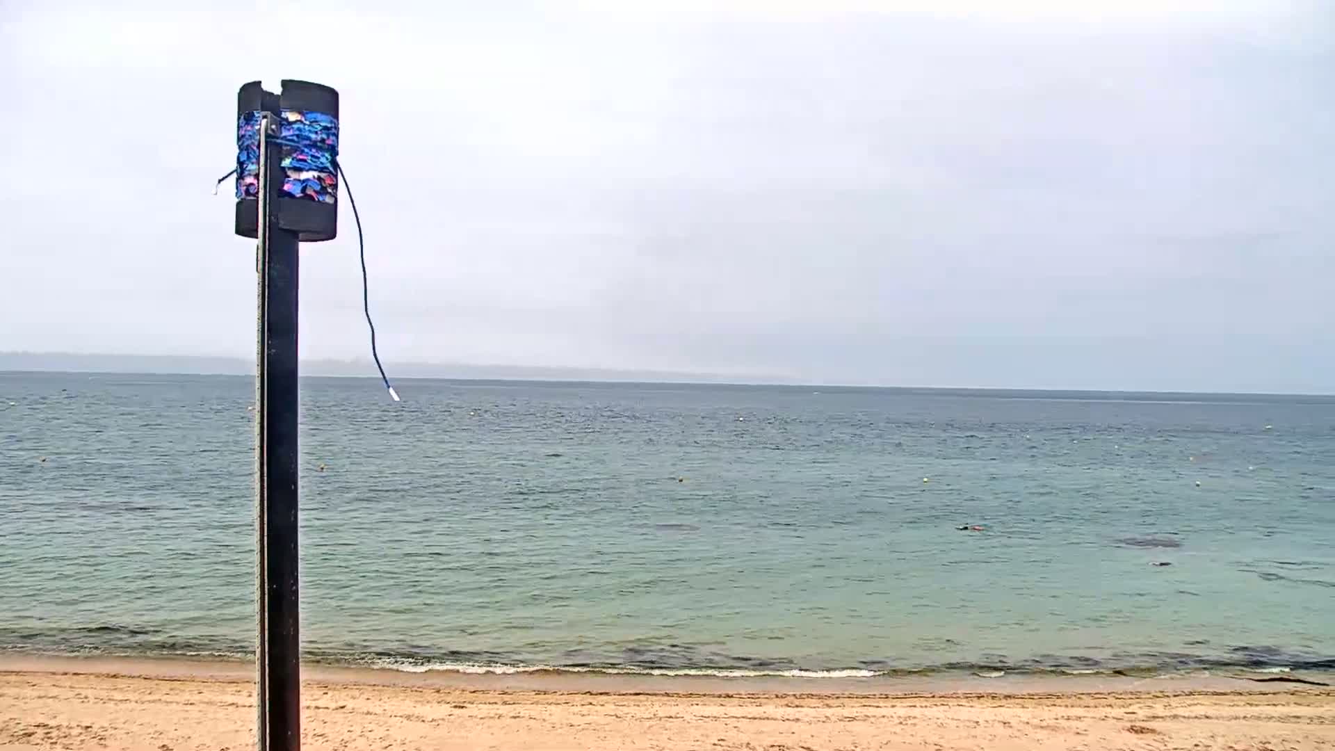 A tall, thin post with colorful fabric wrapped around it stands on a sandy beach overlooking a calm, light-green ocean under an overcast sky.
