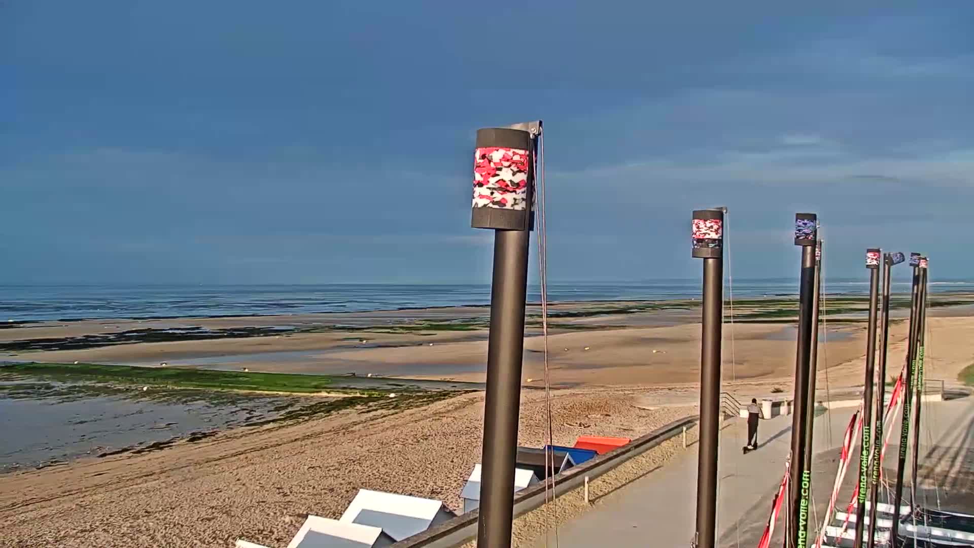 A sandy beach stretches to a calm ocean under a clear blue sky, with several tall poles topped with patterned cylinders situated on a paved area near the shoreline.