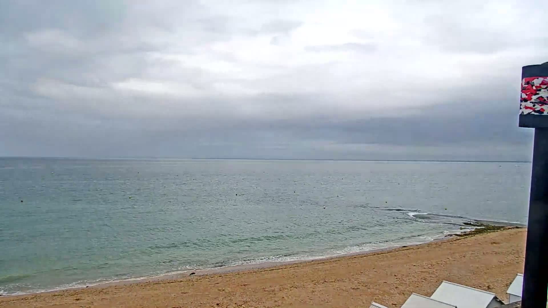 A sandy beach slopes gently down to a calm, light-green sea under a cloudy, overcast sky.