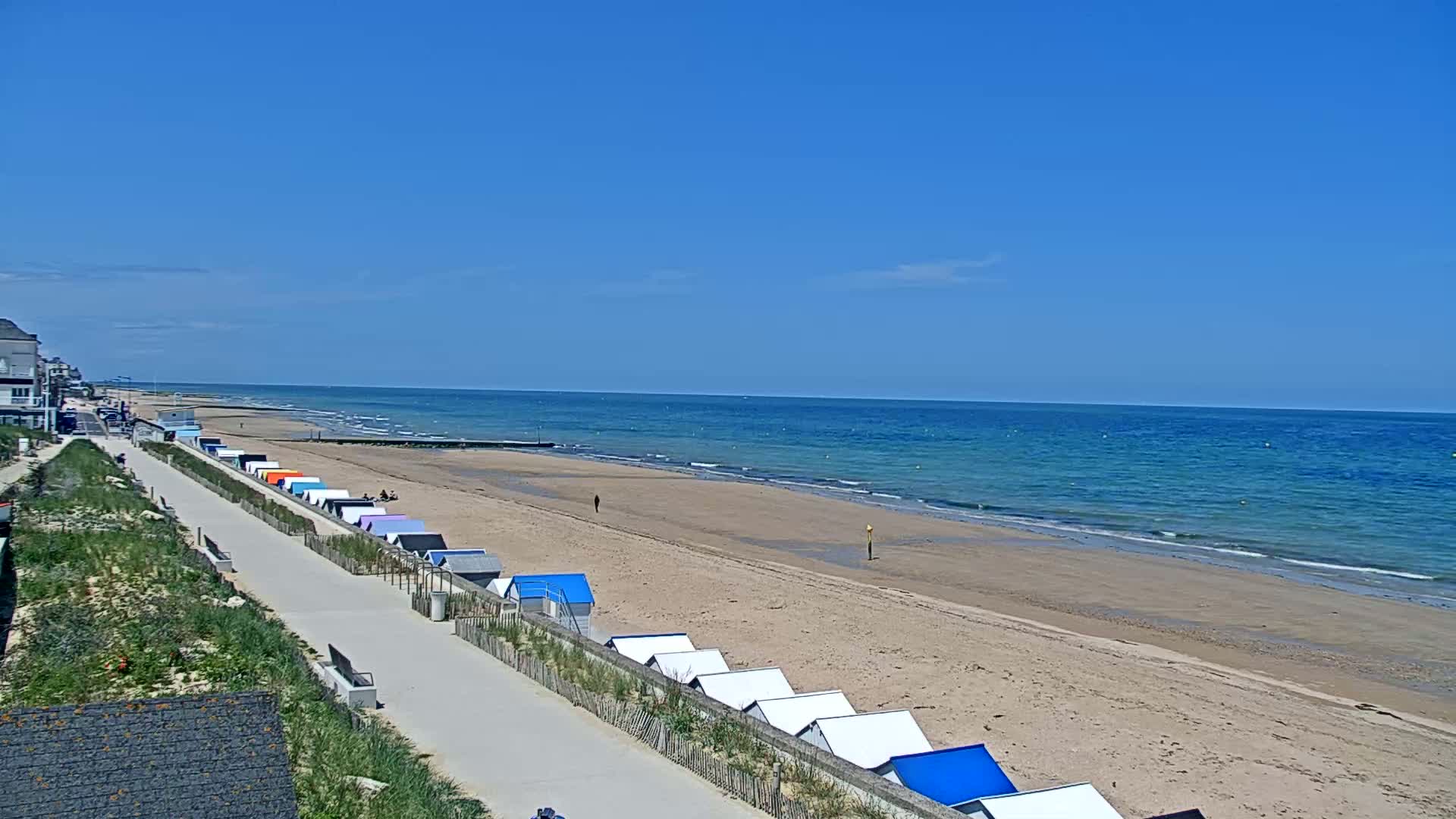 A sandy beach with colorful beach huts lines a paved walkway leading to a calm, blue ocean under a clear blue sky.