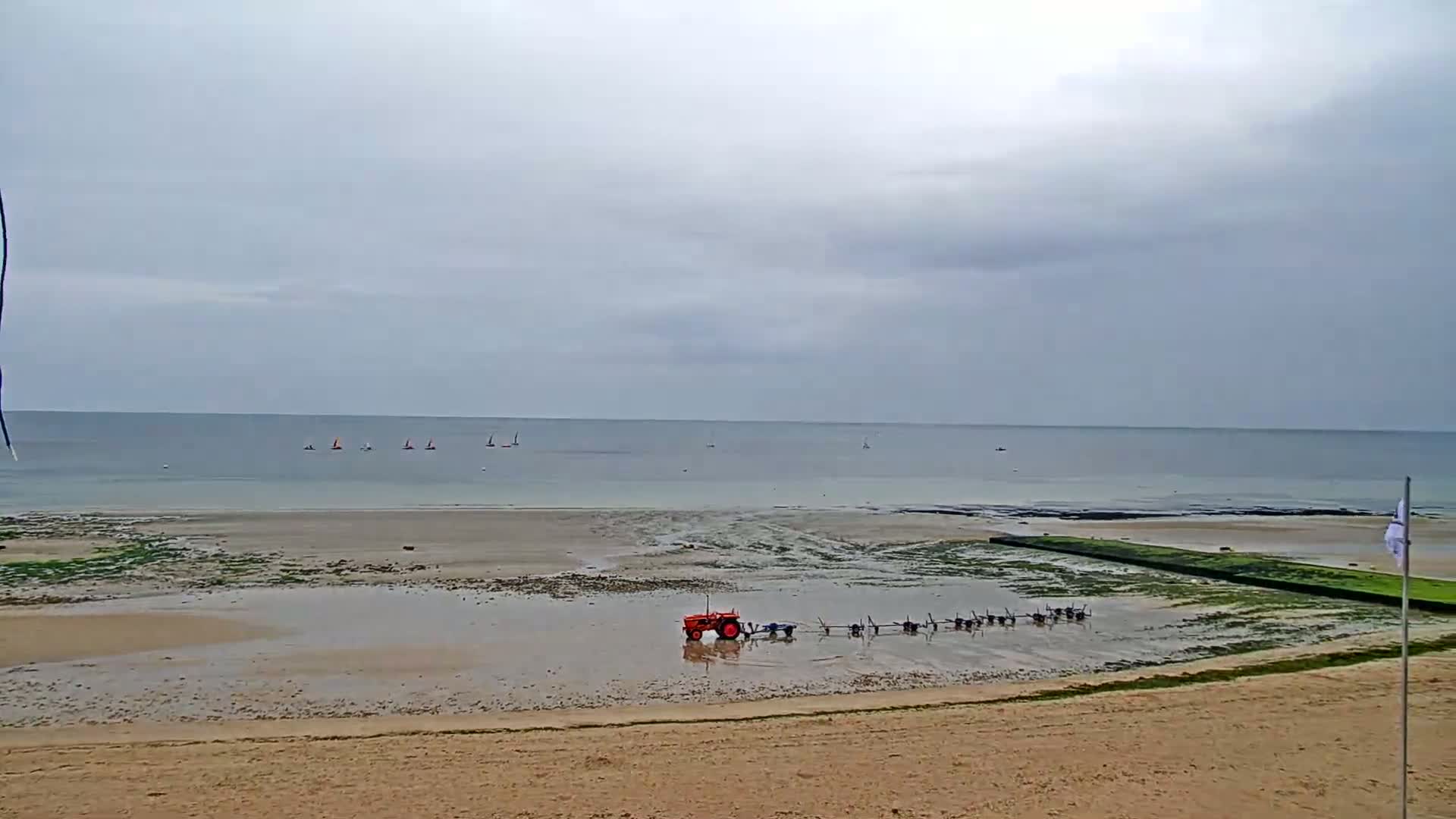 A tractor and several pieces of equipment are on a wet, sandy beach under an overcast sky, with several sailboats visible in the calm sea in the distance.