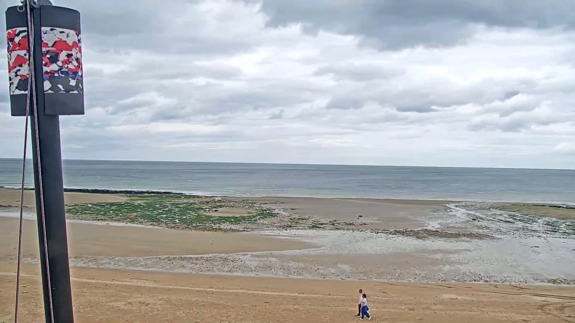 A couple walks on a sandy beach under a cloudy sky, with a calm ocean visible in the background.