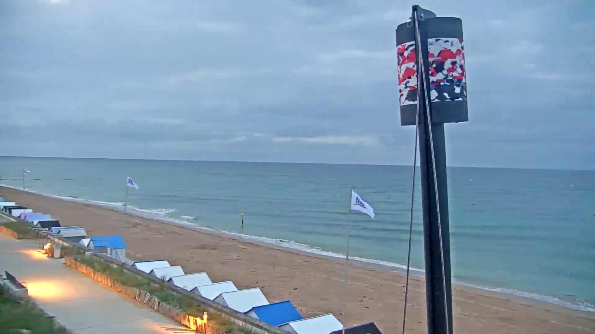 A sandy beach with small beach huts lines a calm, teal ocean under a mostly cloudy sky.