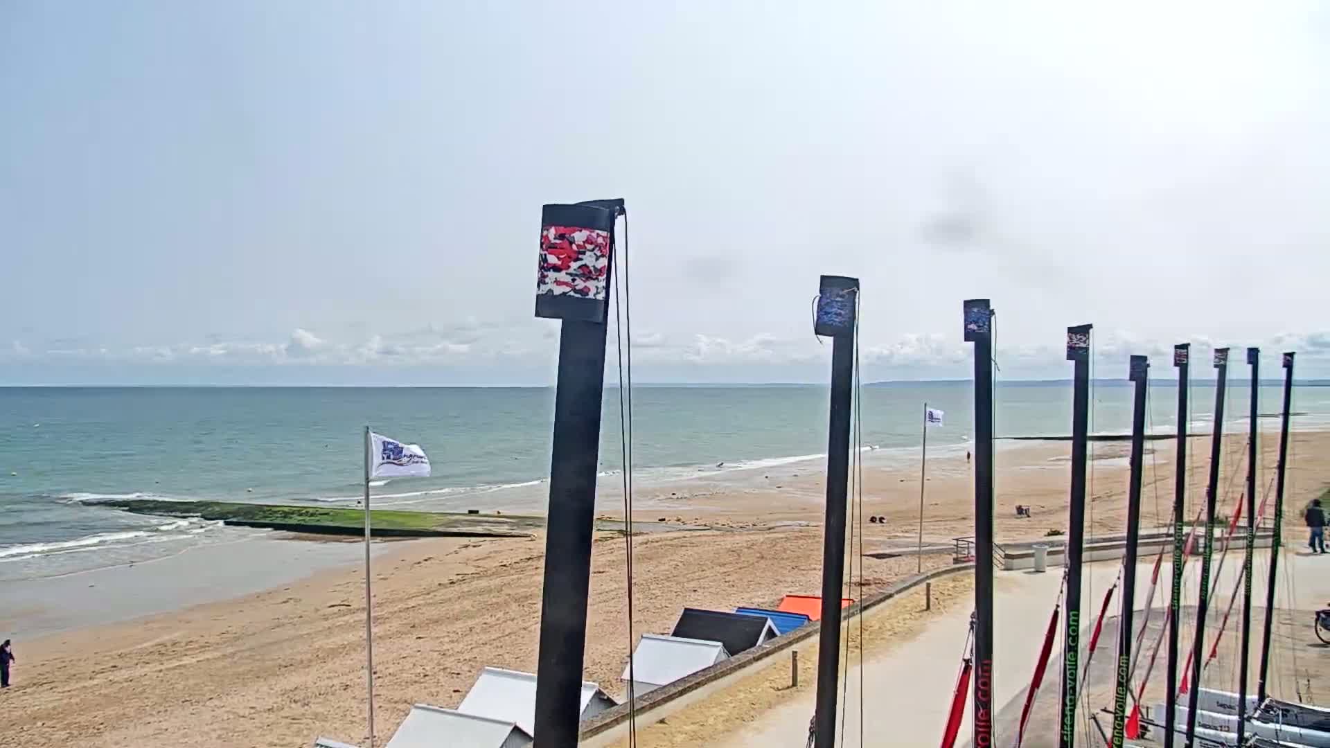 A sandy beach with calm ocean waves under a mostly sunny sky is lined with tall masts, beach huts, and a few people.