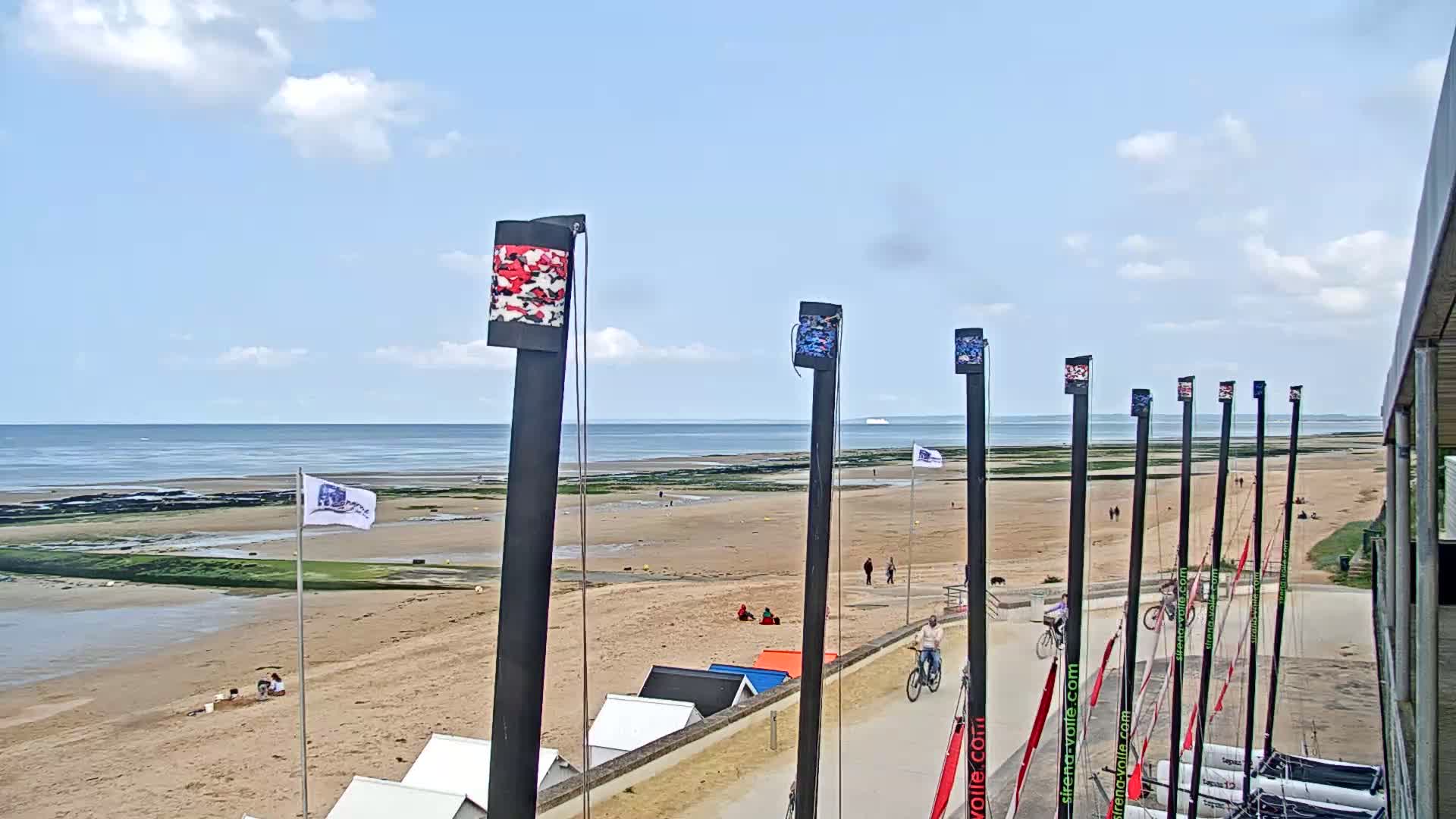 A sandy beach with a calm ocean in the background on a partly cloudy day, and several tall posts with colorful decorations stand along a paved path near beach huts.