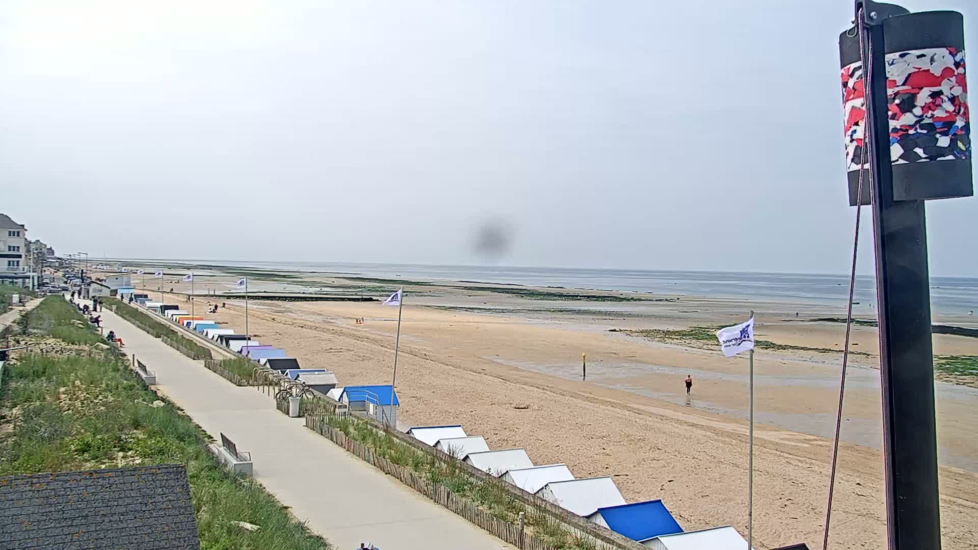 A sandy beach with rows of beach huts stretches alongside a paved walkway leading to buildings, all under an overcast sky.