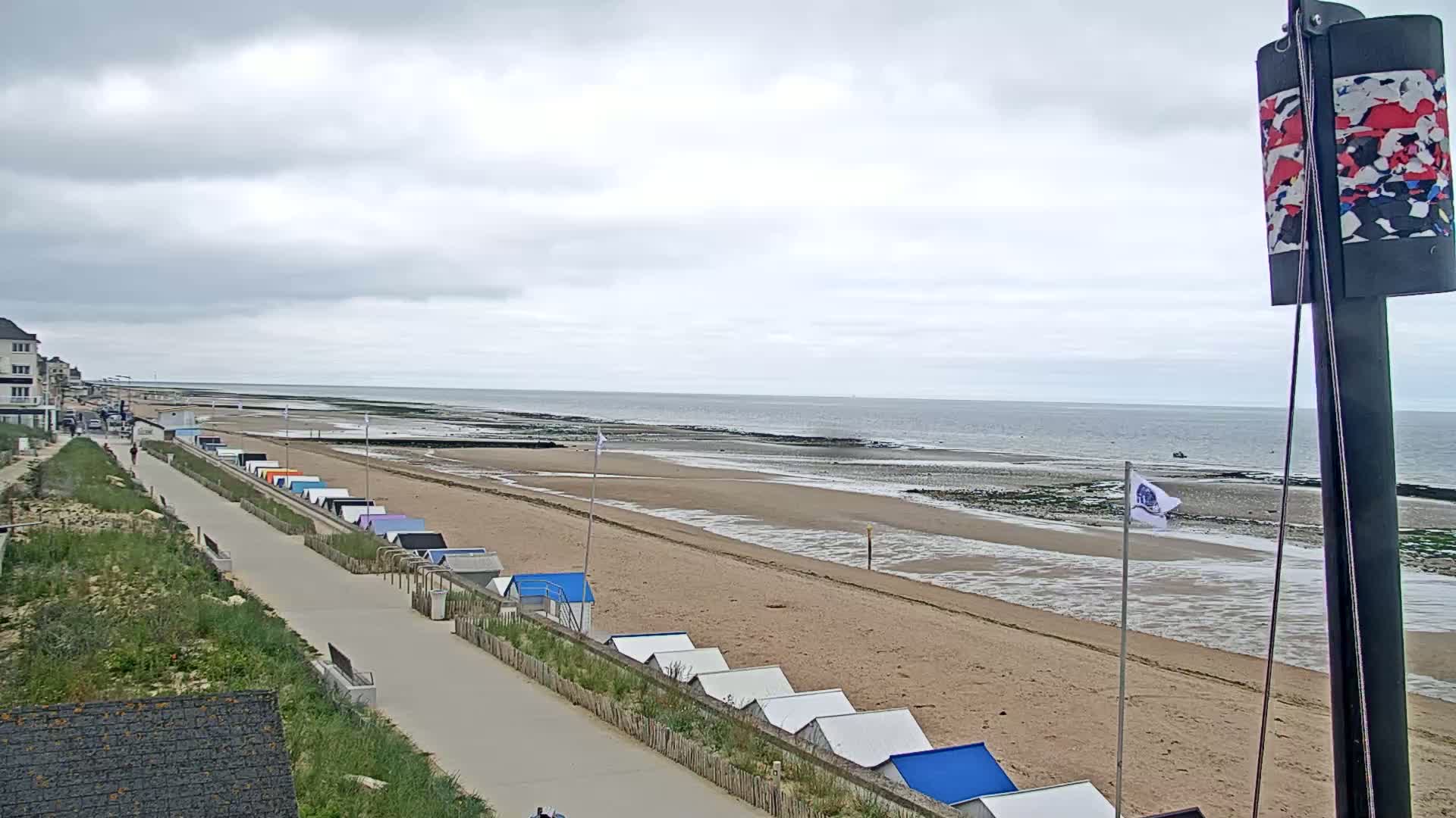 A sandy beach with colorful beach huts lines a paved walkway, under an overcast sky, next to a calm ocean.