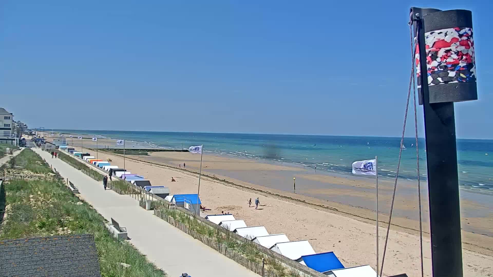 A sandy beach with colorful beach huts and a paved walkway stretches along a calm, turquoise ocean under a clear blue sky.