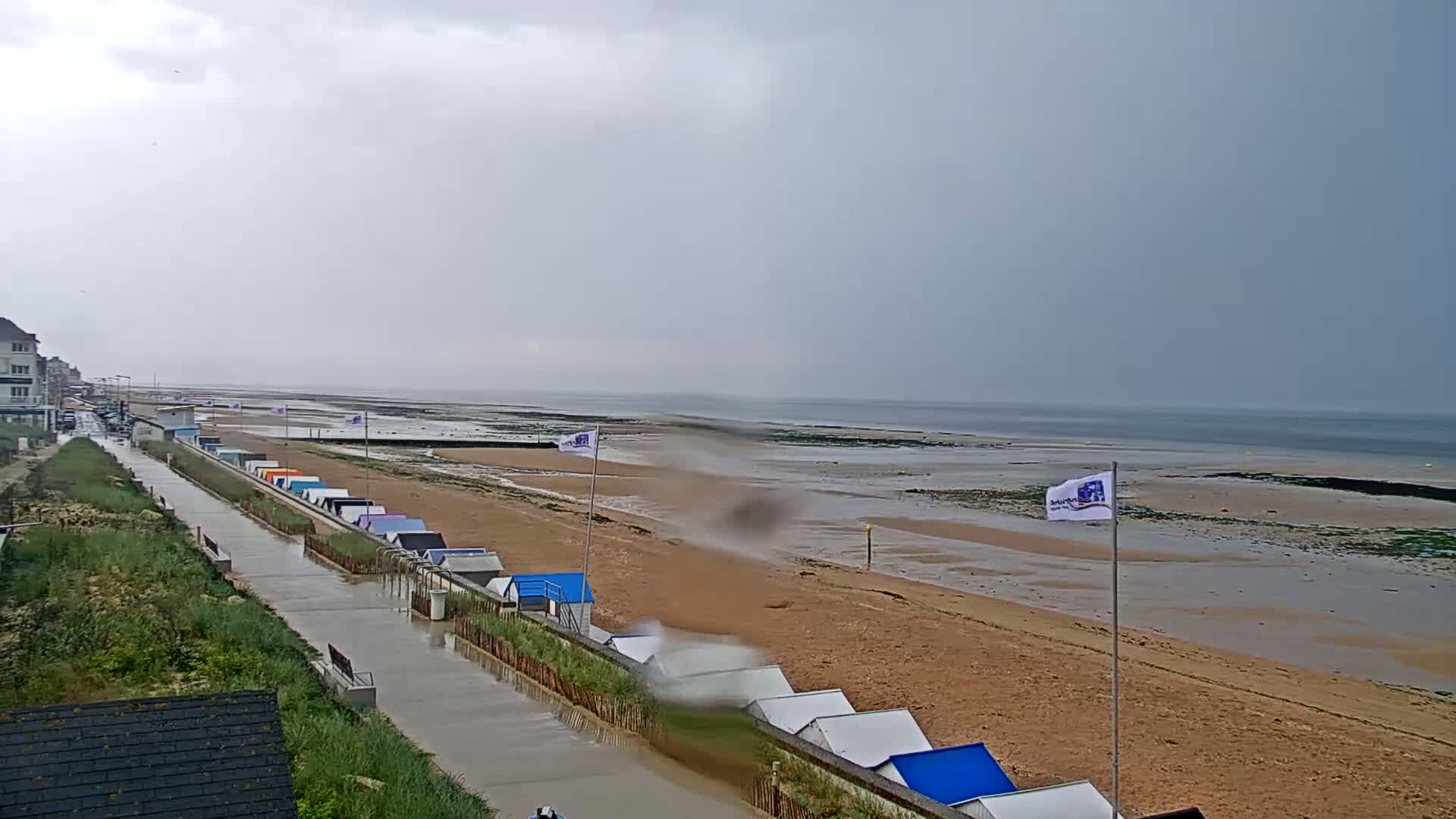 A wet, overcast day shows a sandy beach with rows of small beach huts, a walkway, and buildings in the background.