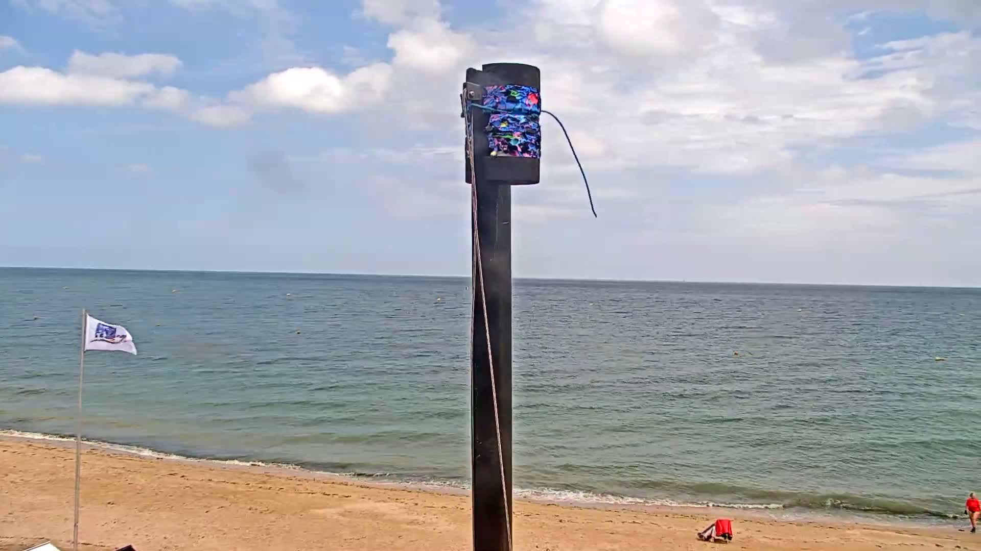 A dark pole with a patterned sleeve attached stands on a sandy beach overlooking a calm, blue-green ocean under a partly cloudy sky.