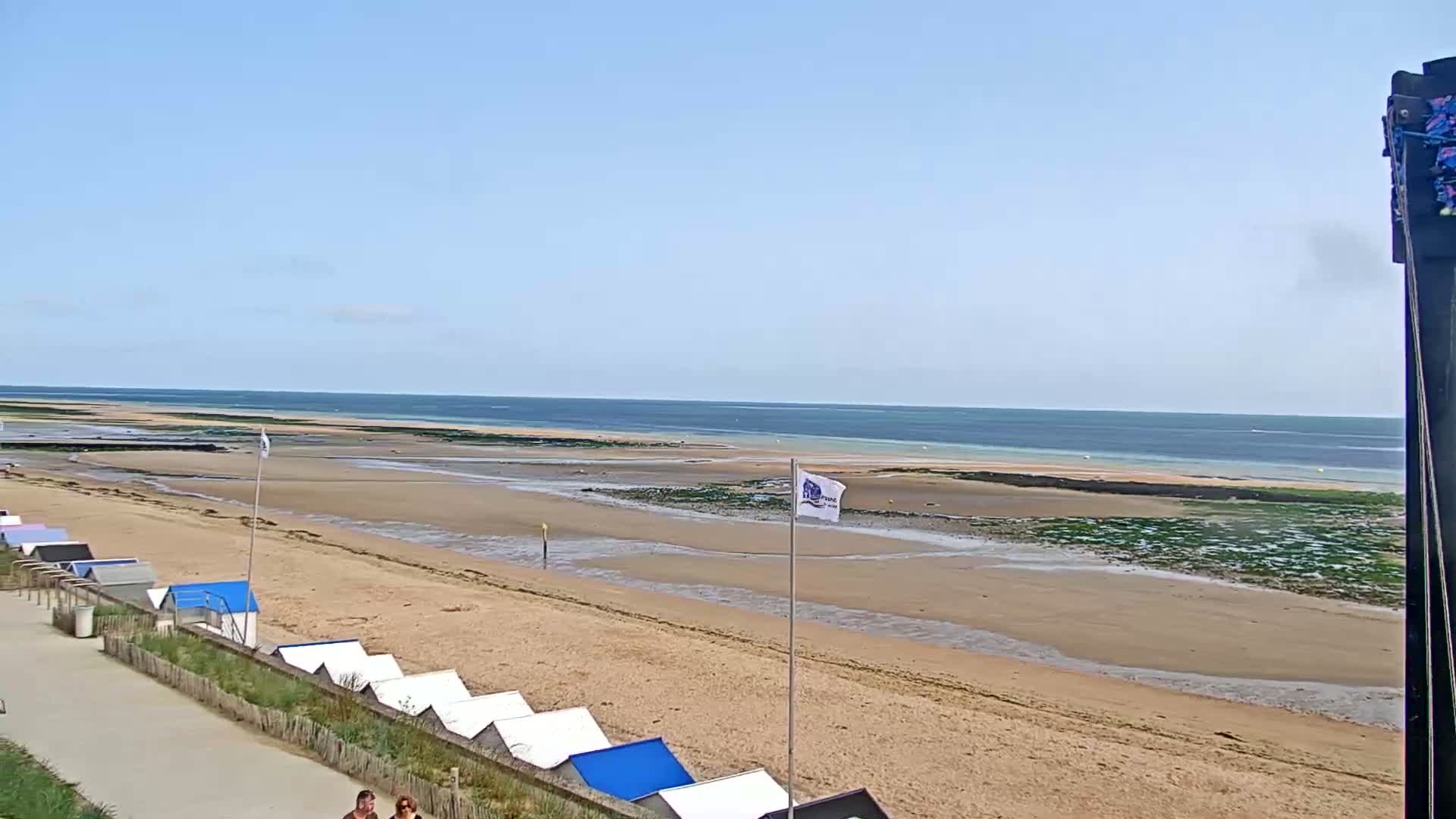 A sandy beach stretches toward a calm ocean under a clear blue sky, with small beach huts lining the shore.
