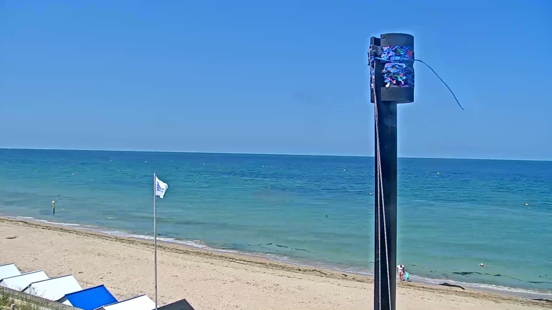 A sandy beach and calm, turquoise ocean are seen under a clear blue sky, with a tall post in the foreground.
