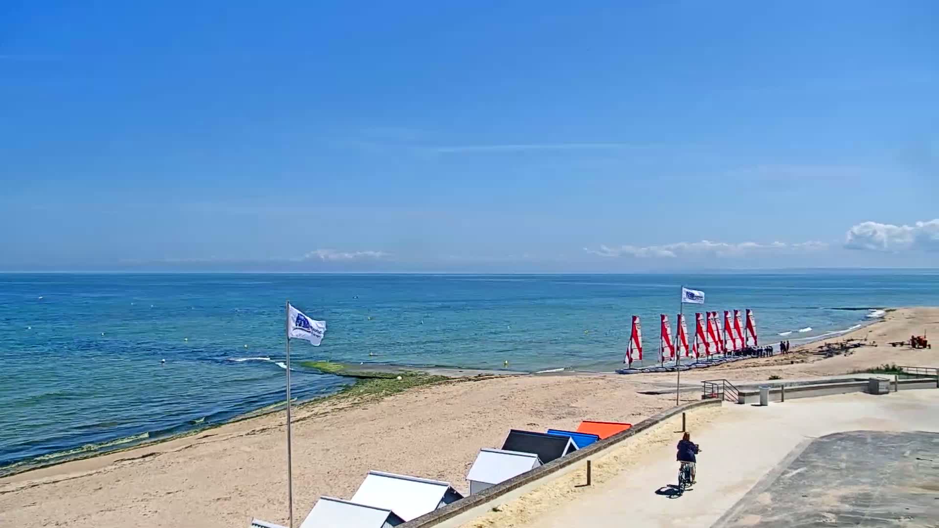 A sandy beach with small beach huts, sailboats on the shore, and a person biking on a paved path overlooks a calm, blue ocean under a mostly sunny sky.
