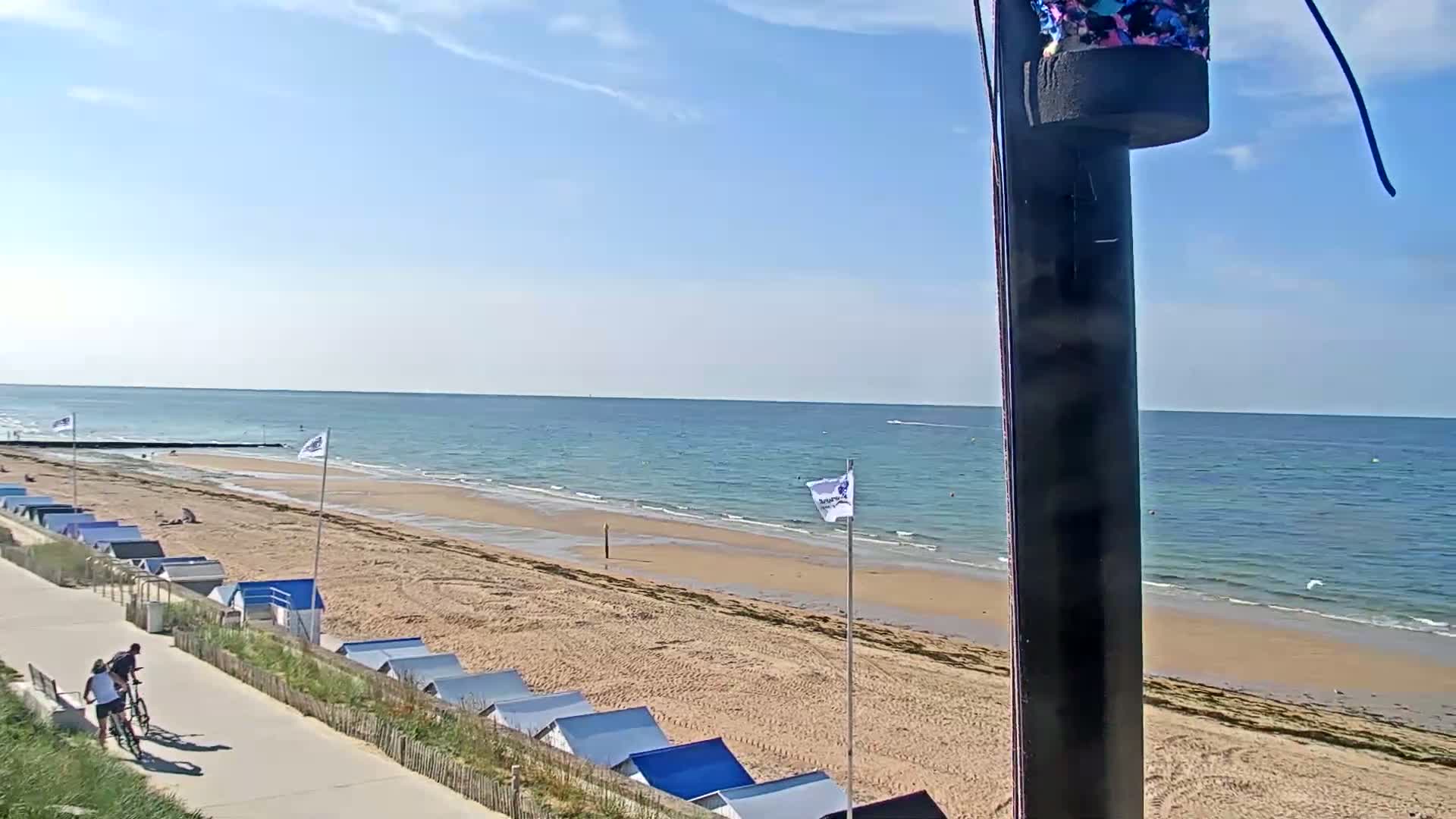 A sandy beach with calm ocean waves under a sunny sky, features small blue beach huts and two people biking along a paved path.