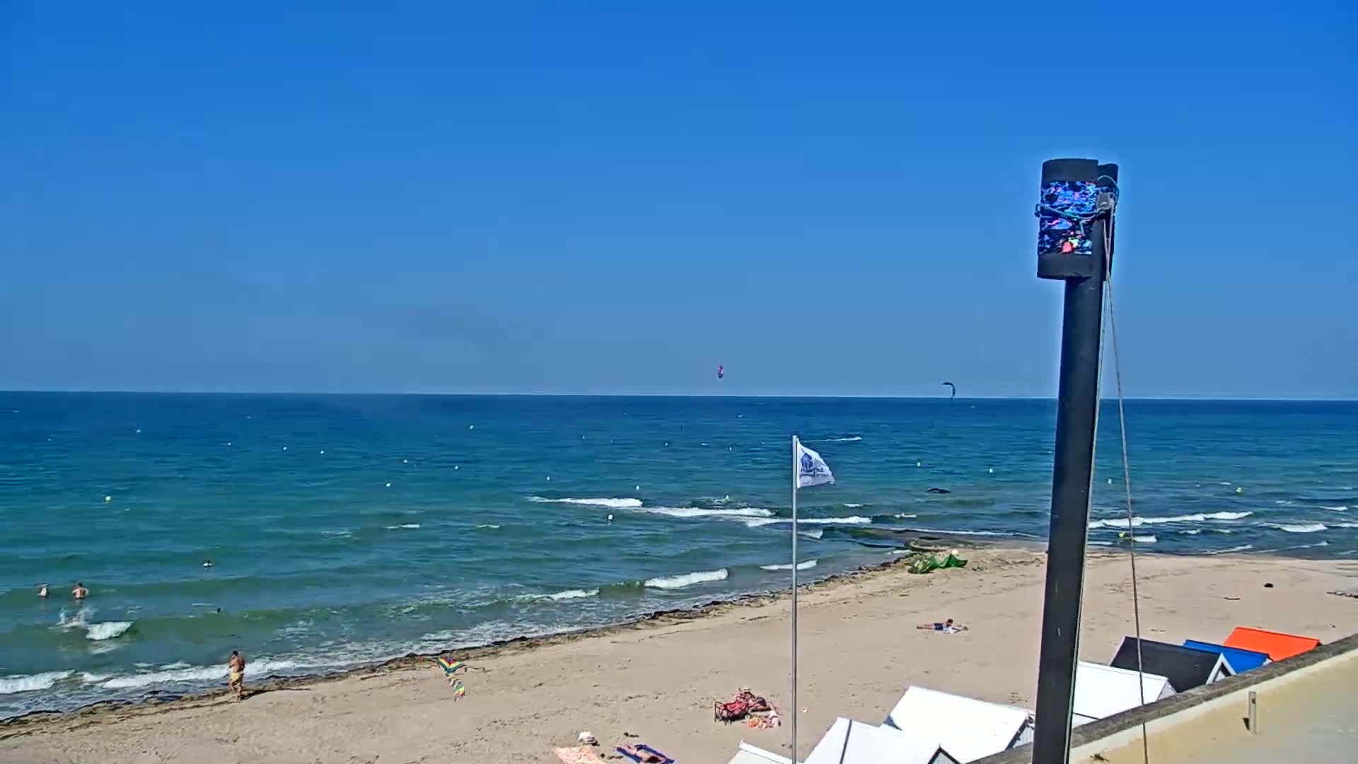 A sandy beach with people sunbathing and swimming, under a clear blue sky, with a calm teal sea and two kitesurfers visible in the distance.