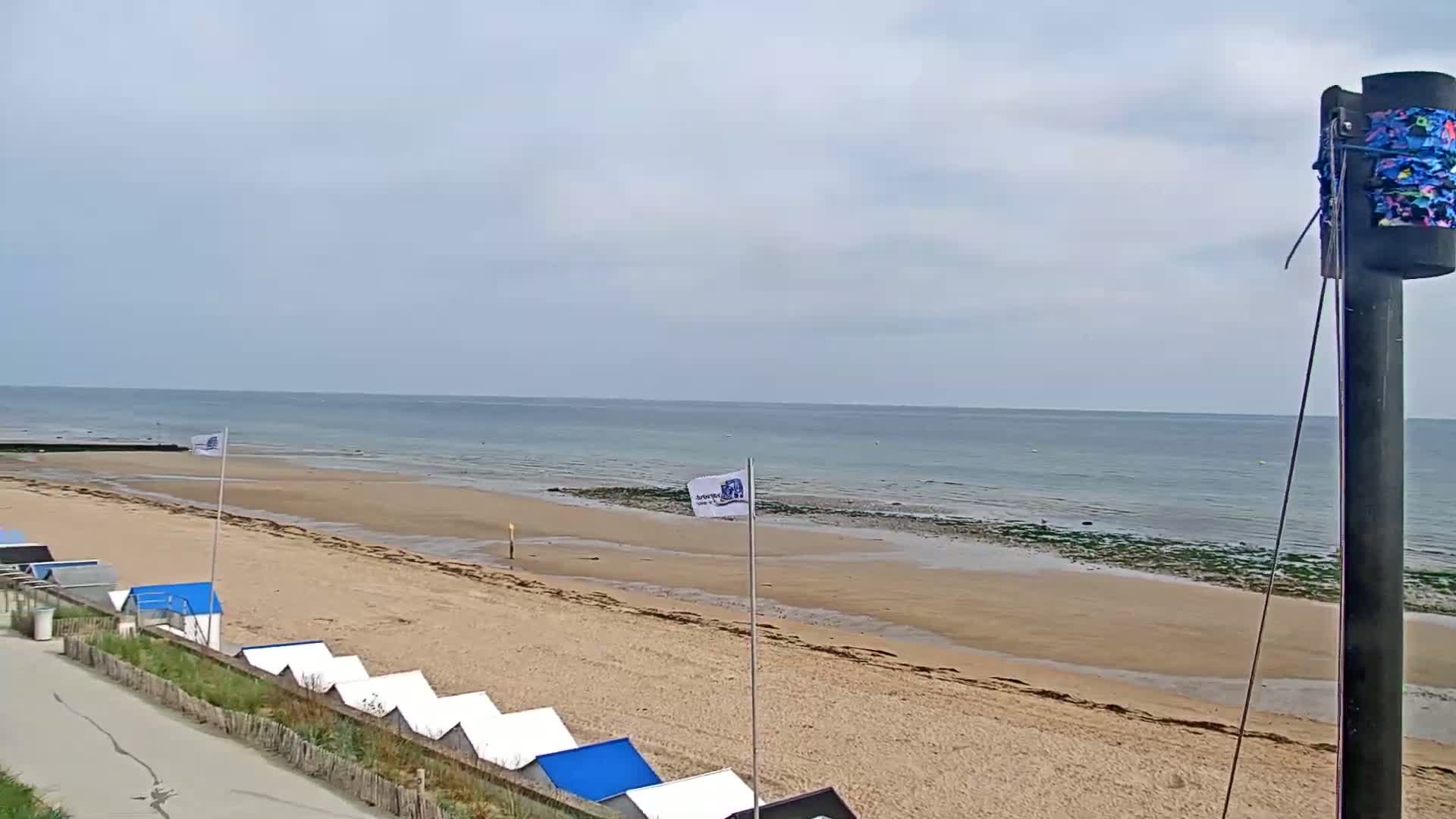 A sandy beach with rows of beach huts stretches toward a calm ocean under an overcast sky.